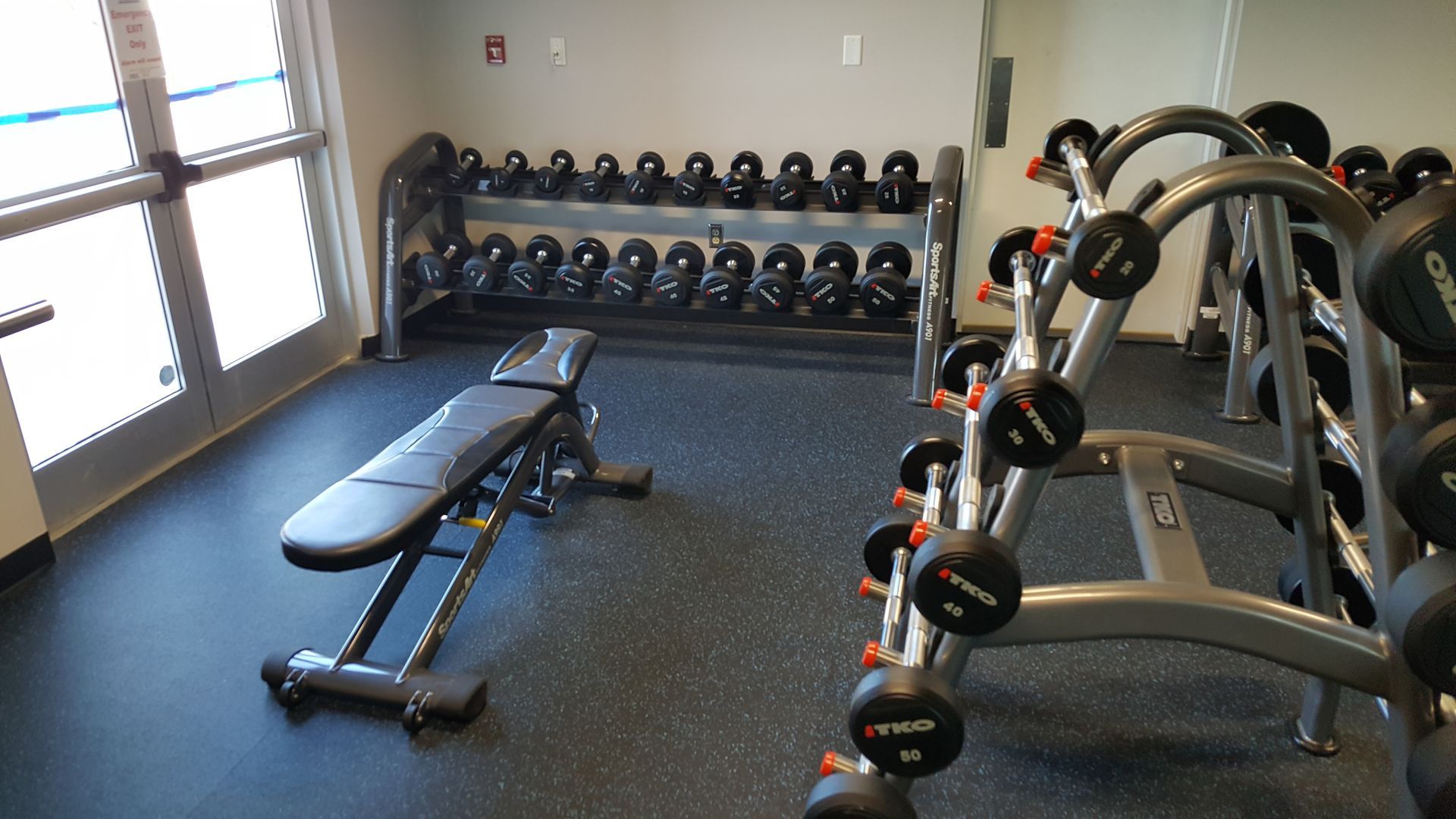 Small gym with dumbbell rack, weight bench, and cable machines on rubber flooring near windows.
