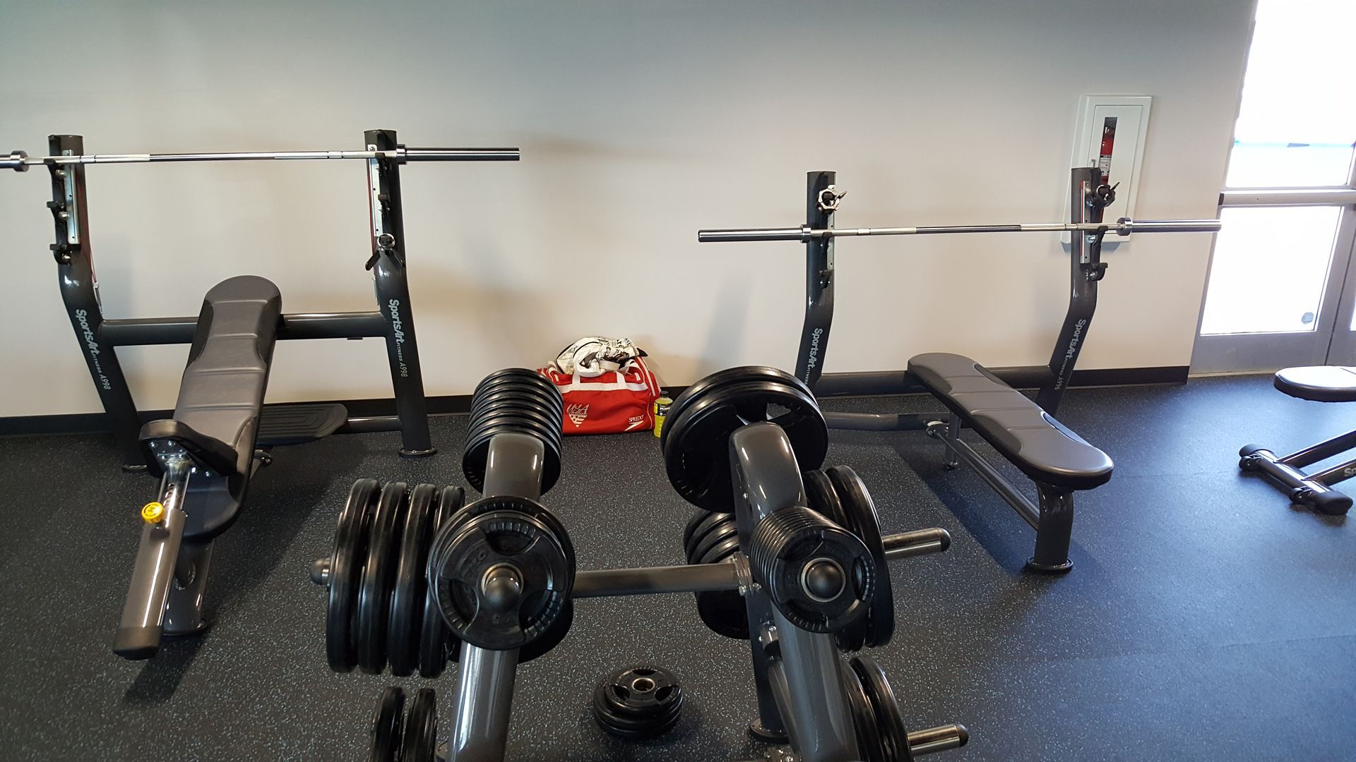 Gym with weight benches, barbells, dumbbells, and a red cleaning cart against a white wall.