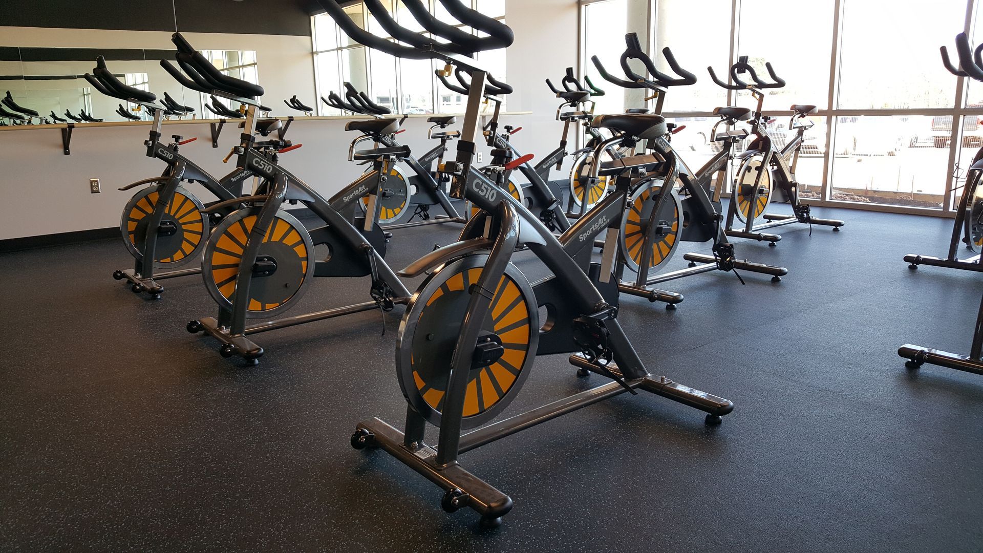 Row of stationary exercise bikes in a bright indoor gym with rubber flooring and large windows
