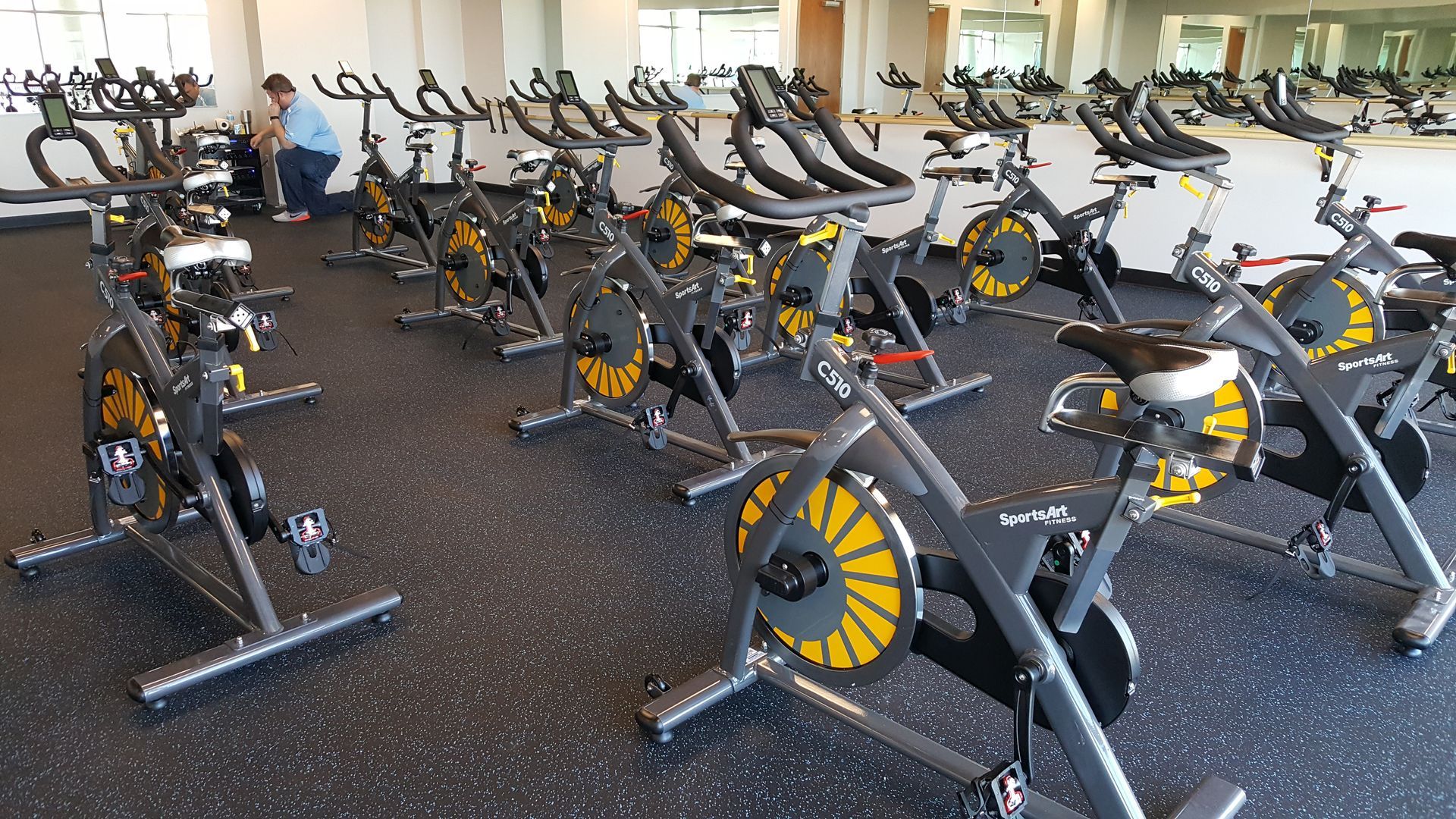Rows of stationary exercise bikes in a bright gym, with gray frames and yellow flywheels.