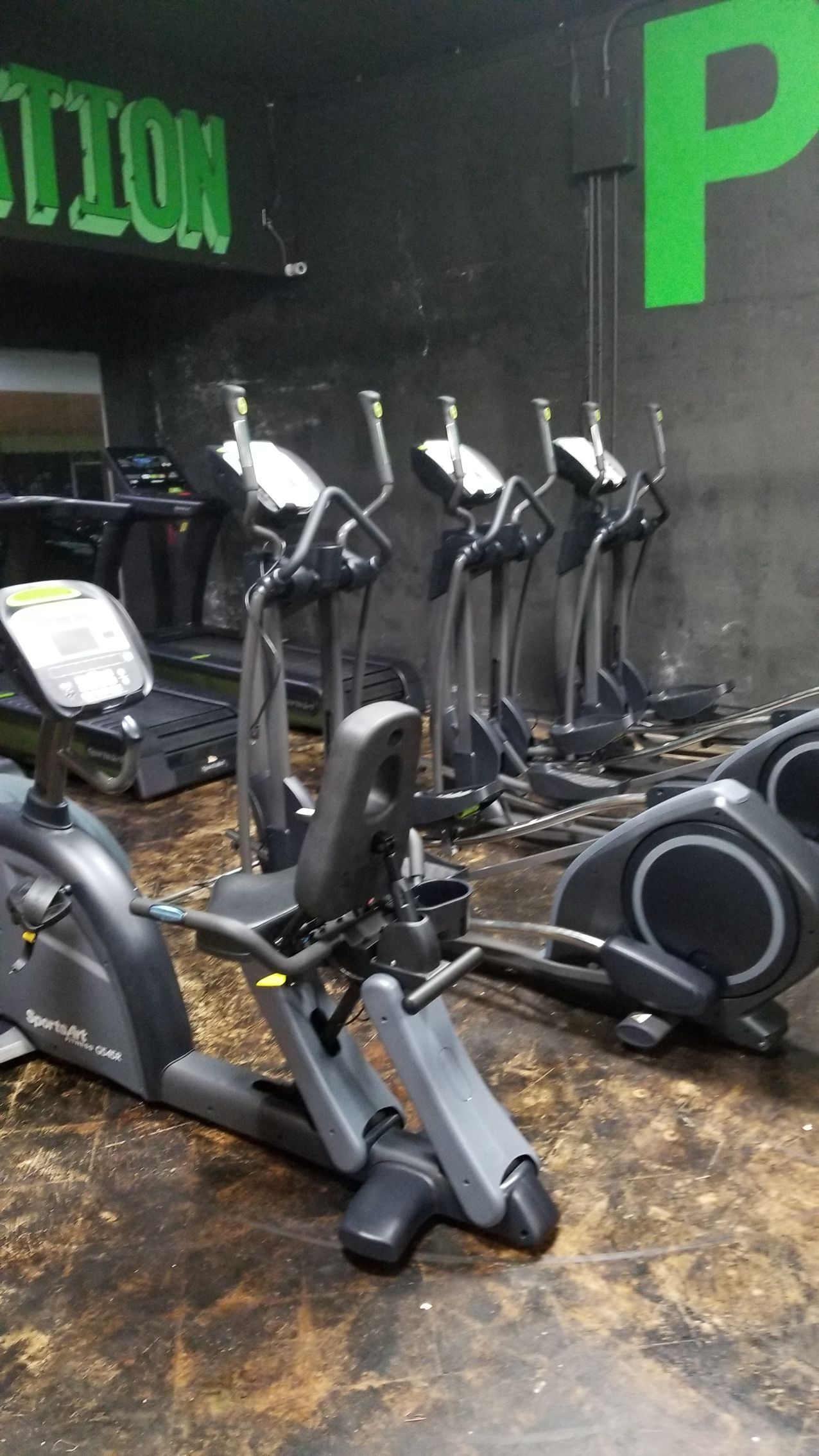 Indoor gym with rows of black elliptical machines on a worn floor against a dark wall