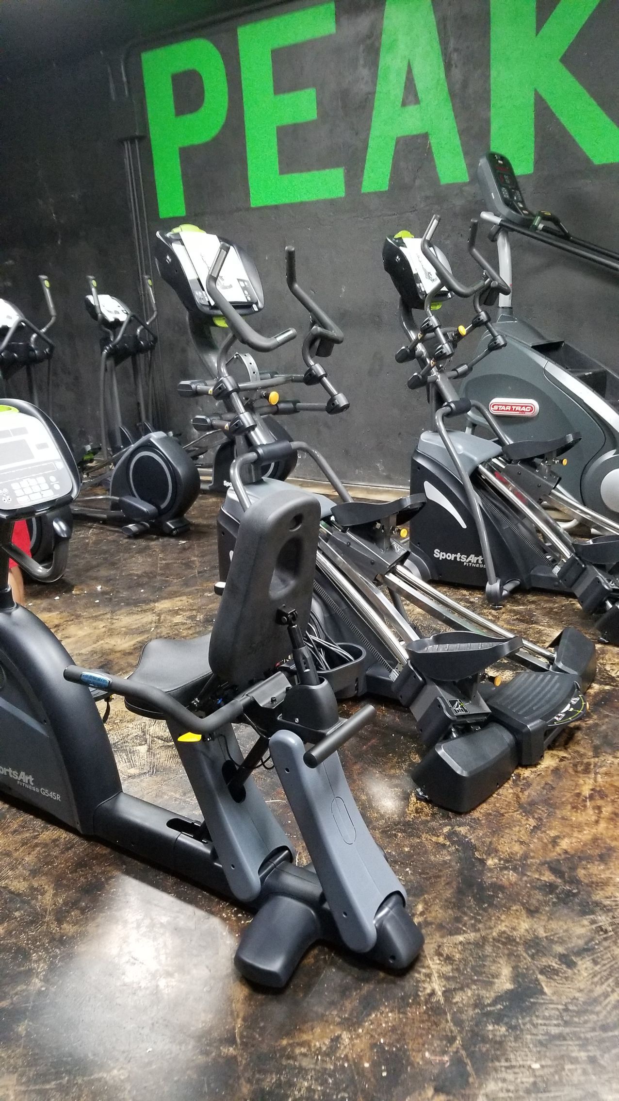 Row of black exercise bikes in a gym, with a green PEAK sign on the wall.
