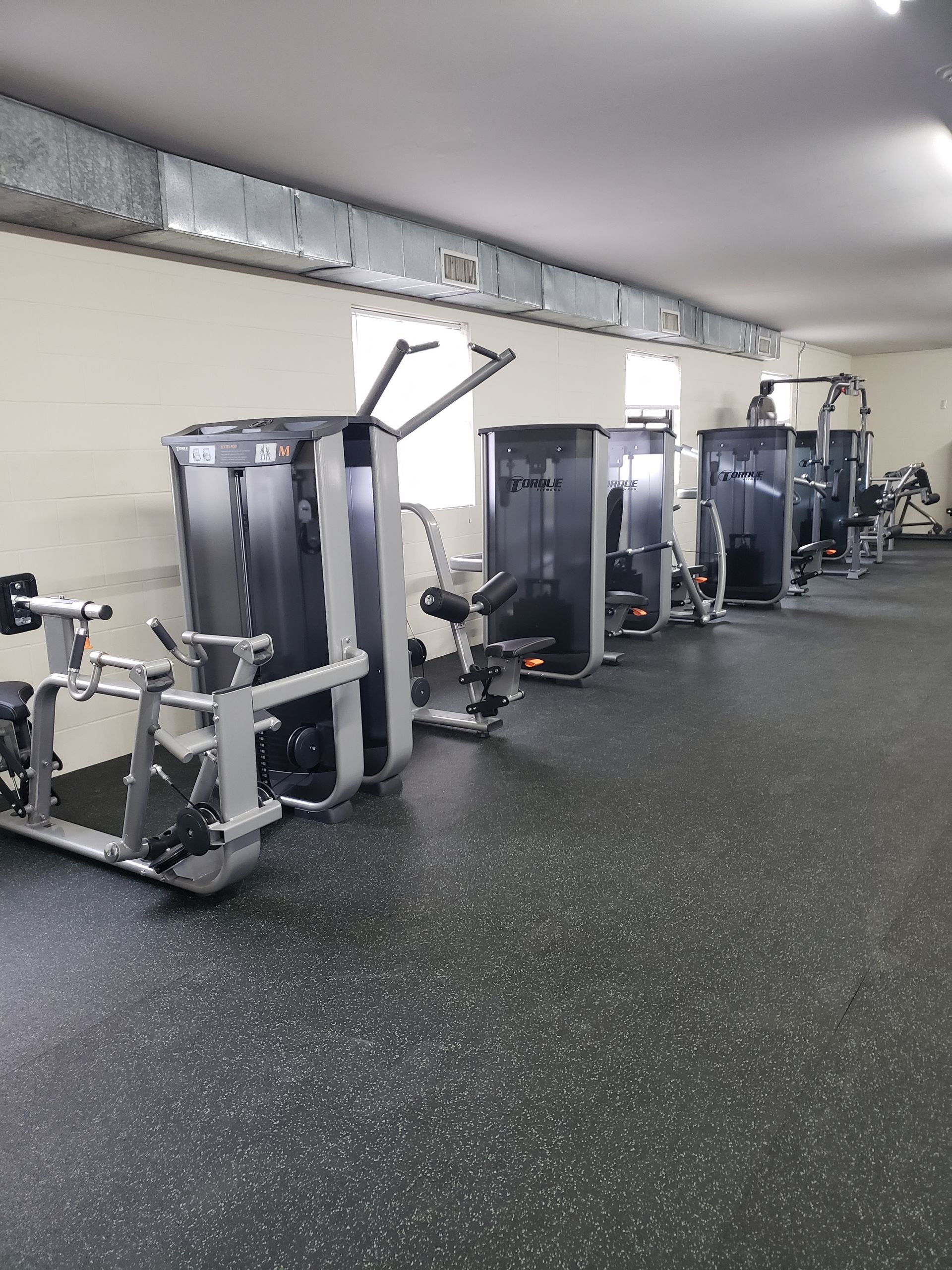 Gym room with rows of strength machines on a rubber floor and mirrors along the wall