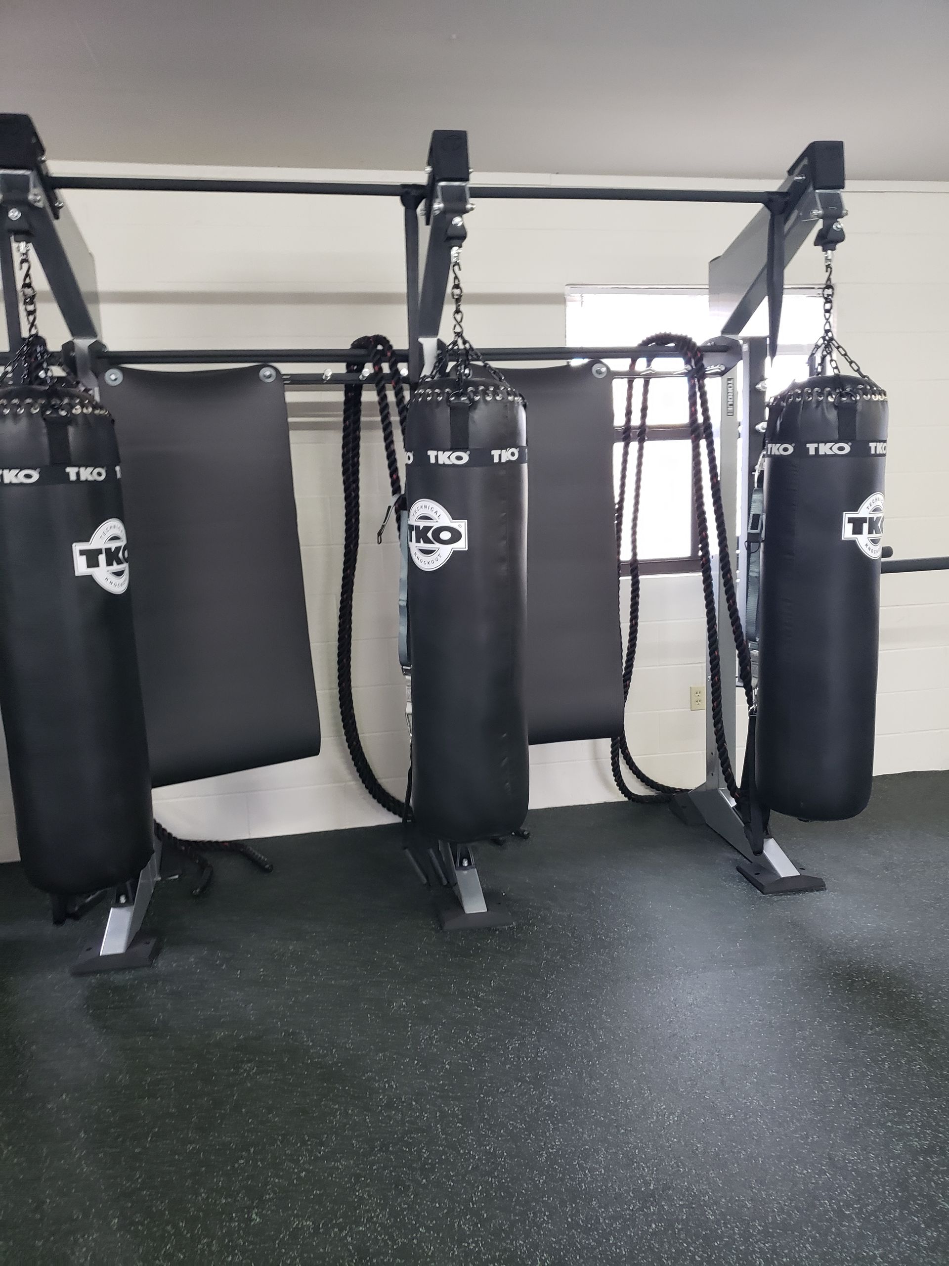 Black hanging punching bags in a gym, with a floor mat and metal support frame.