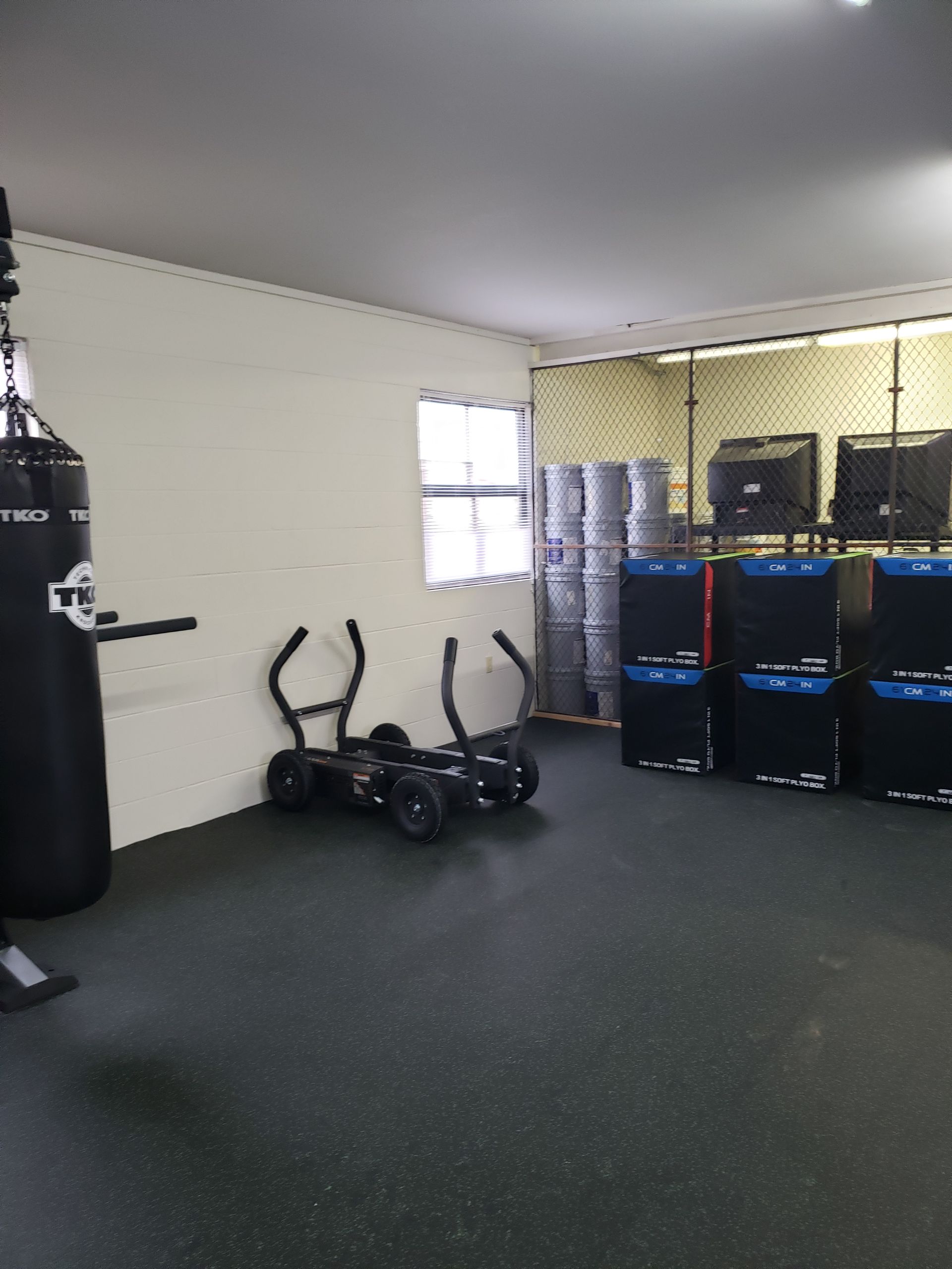 Small gym with punching bag, exercise bikes, stacked weights, and blue storage bins on rubber flooring.
