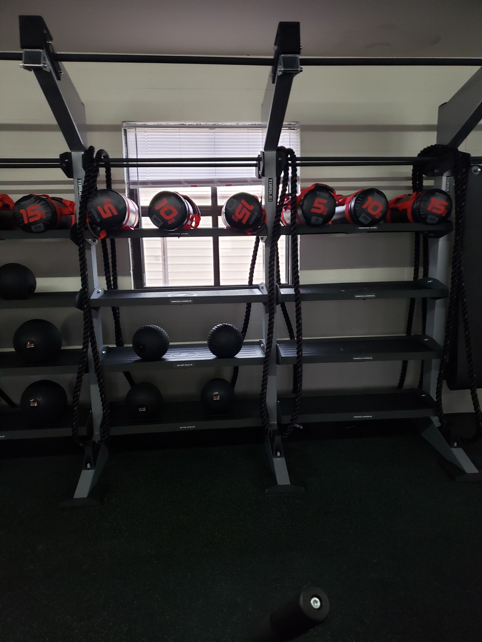 Gym storage shelves with medicine balls and kettlebells in front of a pull-up rack, window behind