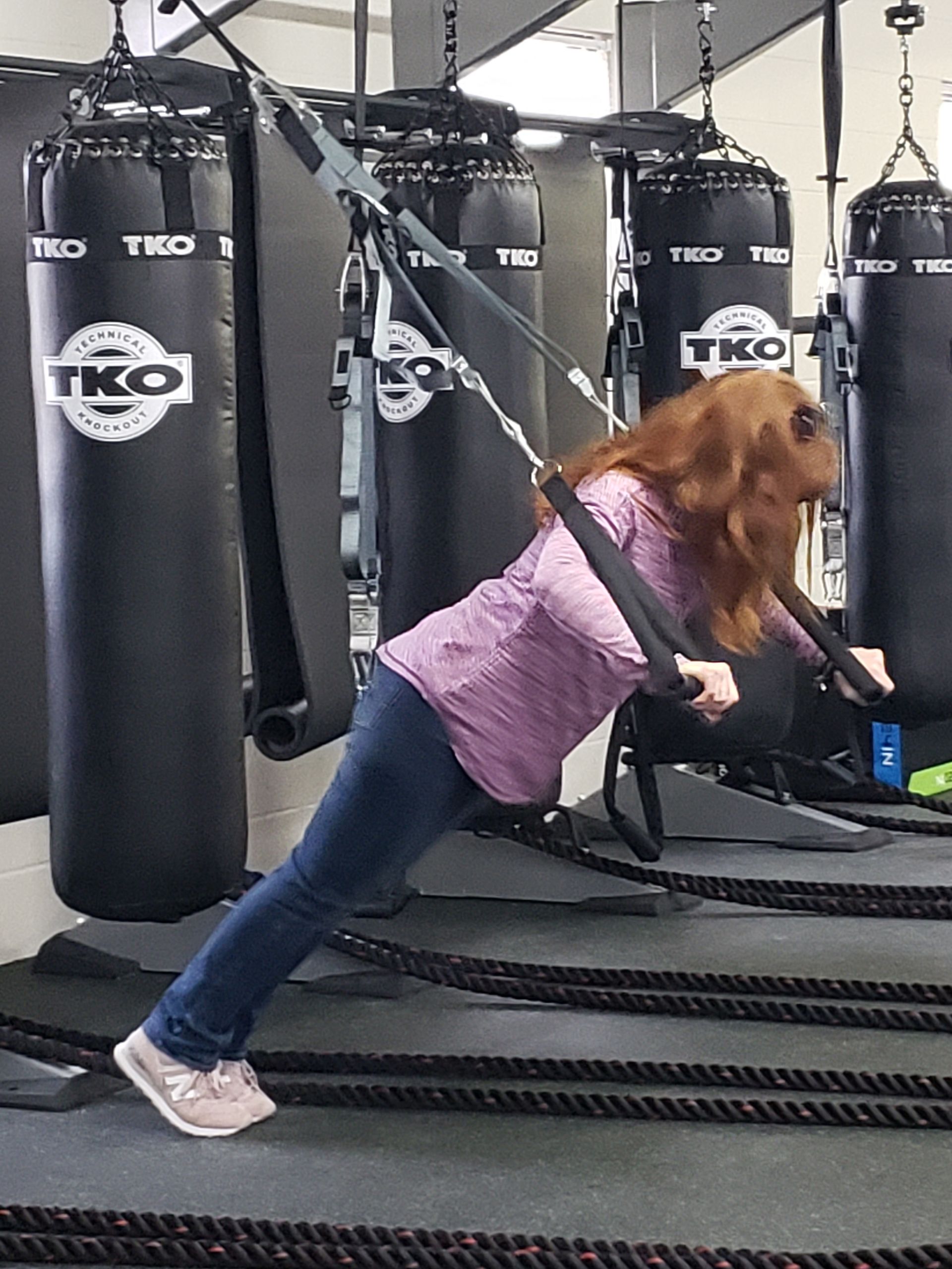 Woman doing incline push-ups with suspension straps in a gym, beside heavy punching bags.
