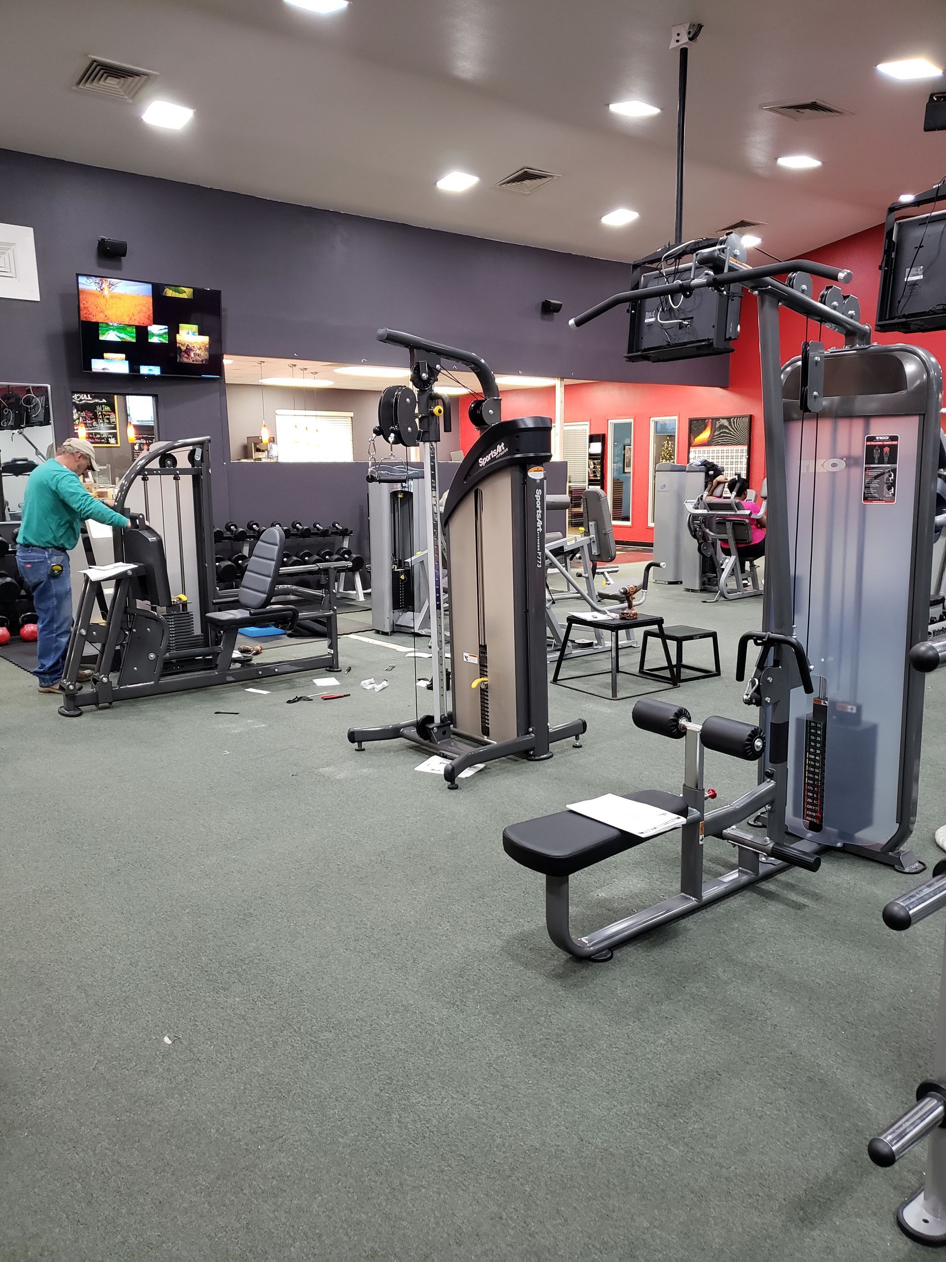 Gym interior with weight machines, benches, and a person using equipment in the background