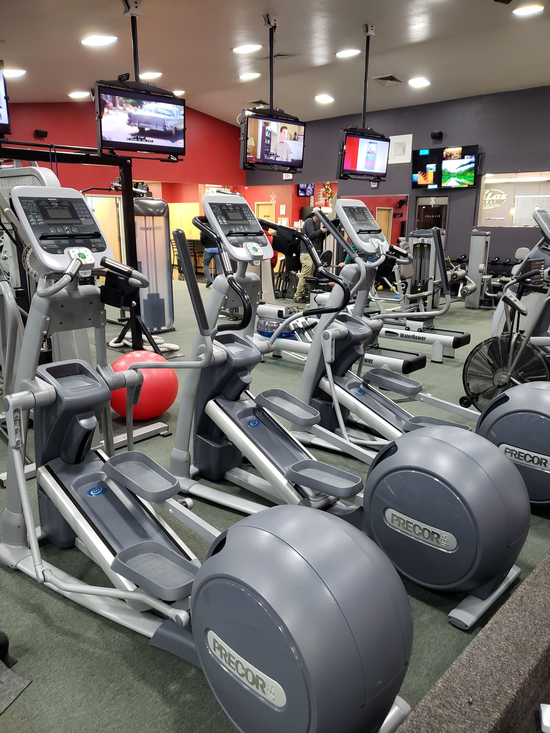 Gym with rows of ellipticals and TVs on a red wall