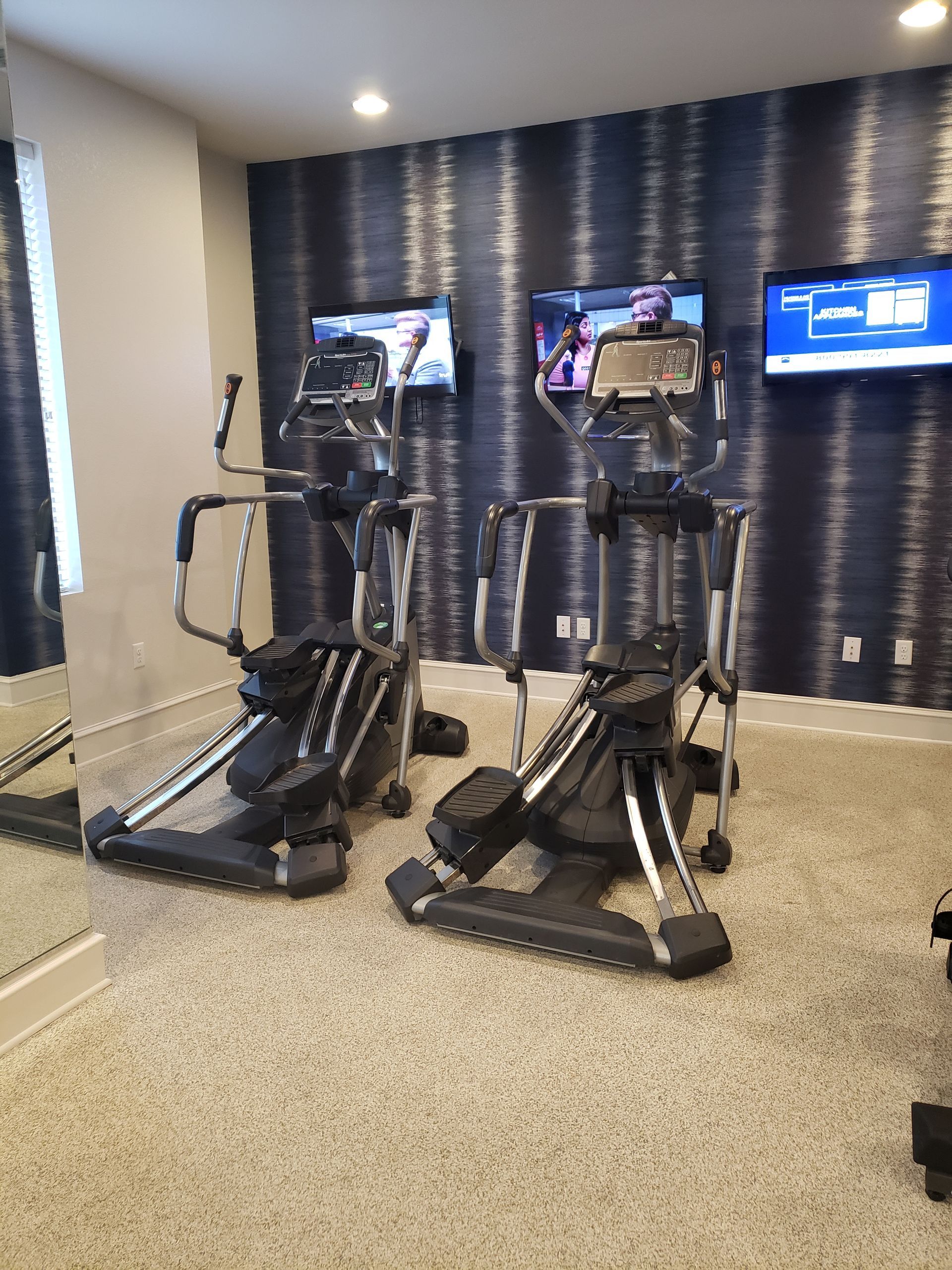 Two black elliptical machines in a gym with wall-mounted TVs and a mirror
