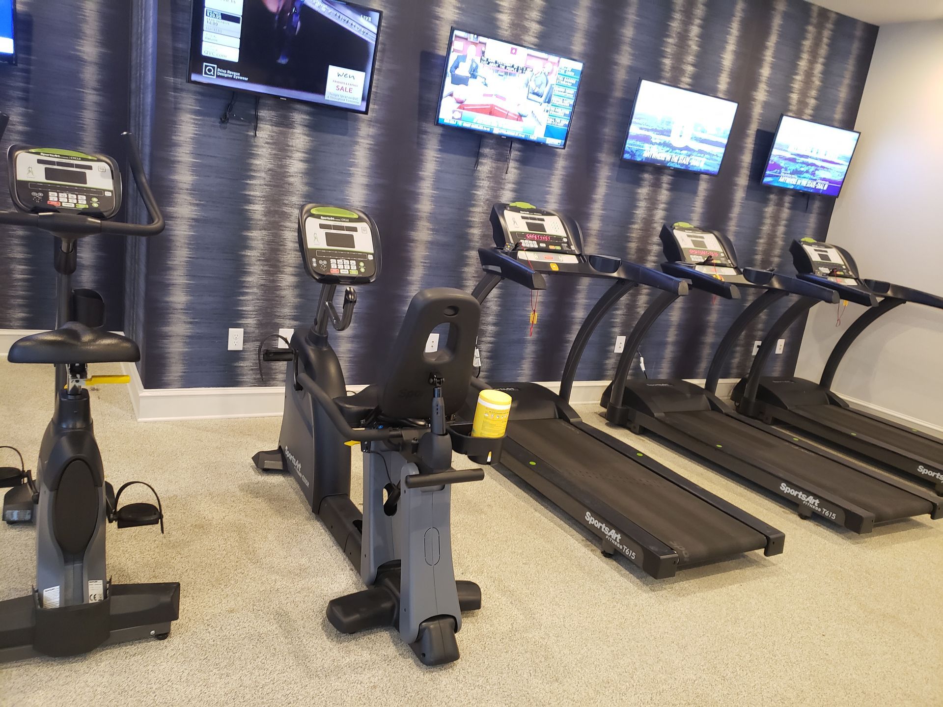 Gym with stationary bikes and treadmills in front of wall-mounted TVs