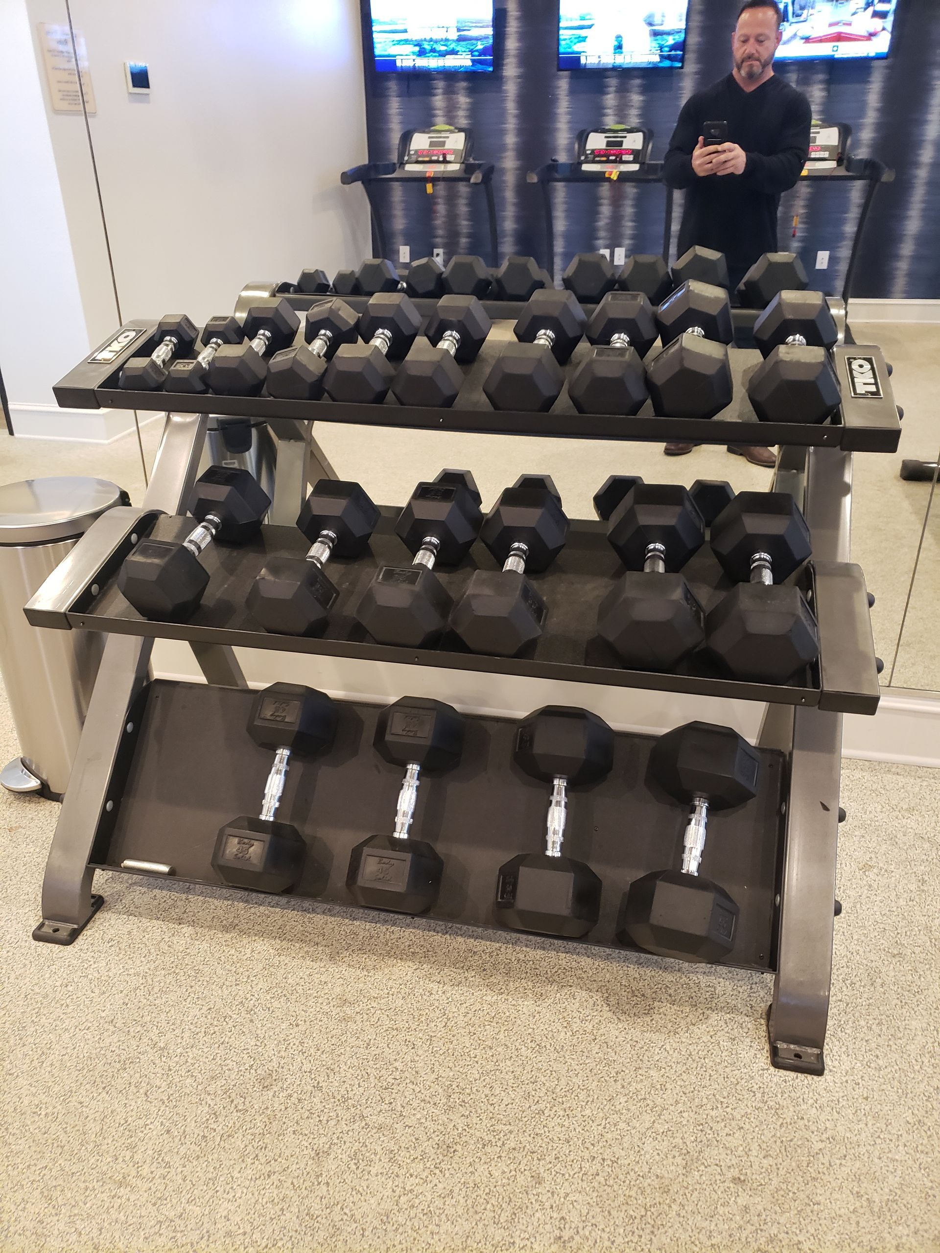 Three-tier rack of black dumbbells in a gym, with a man standing in the background.