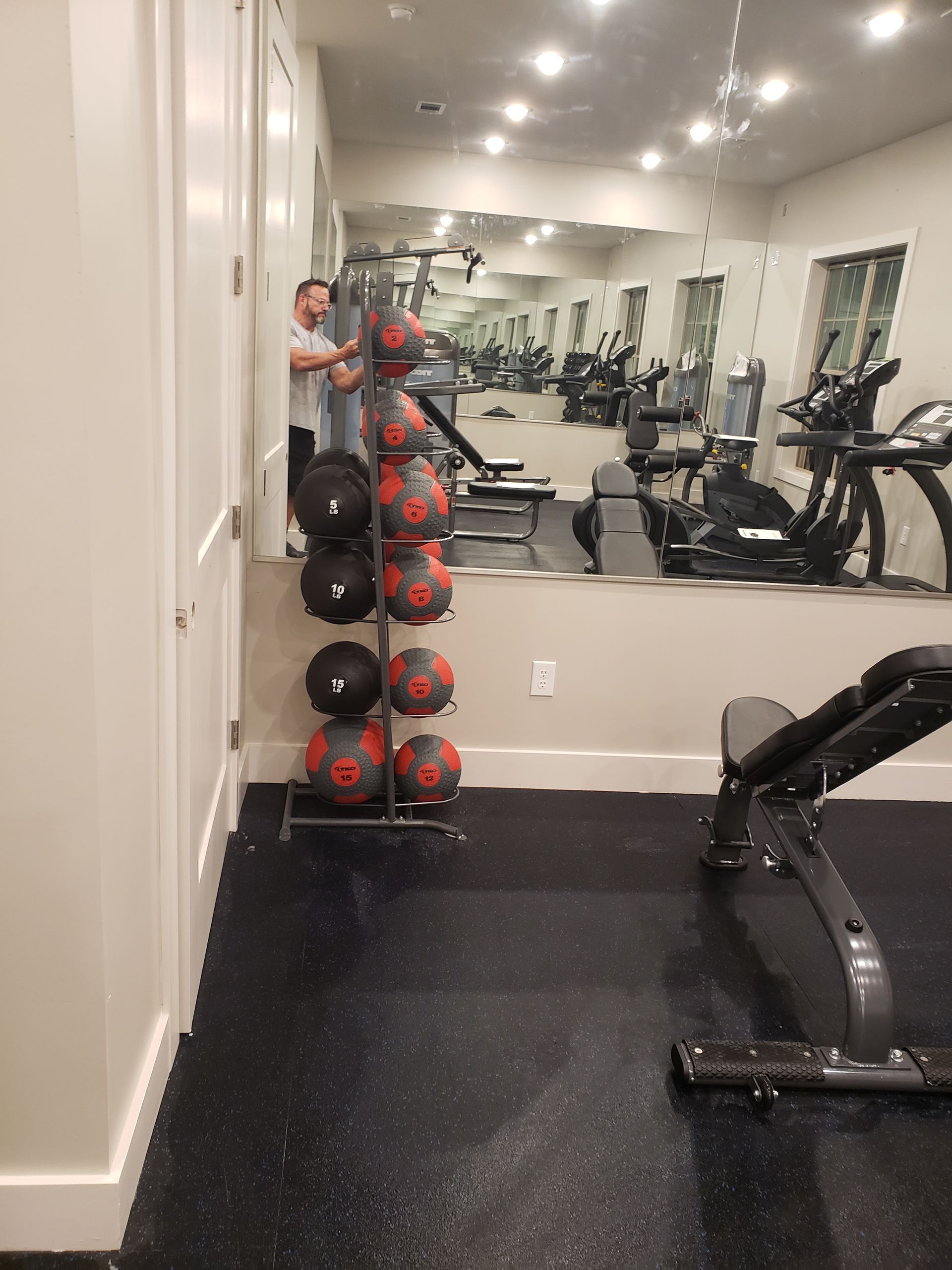 Gym room with mirrored wall, dumbbell rack, and exercise bike on black flooring