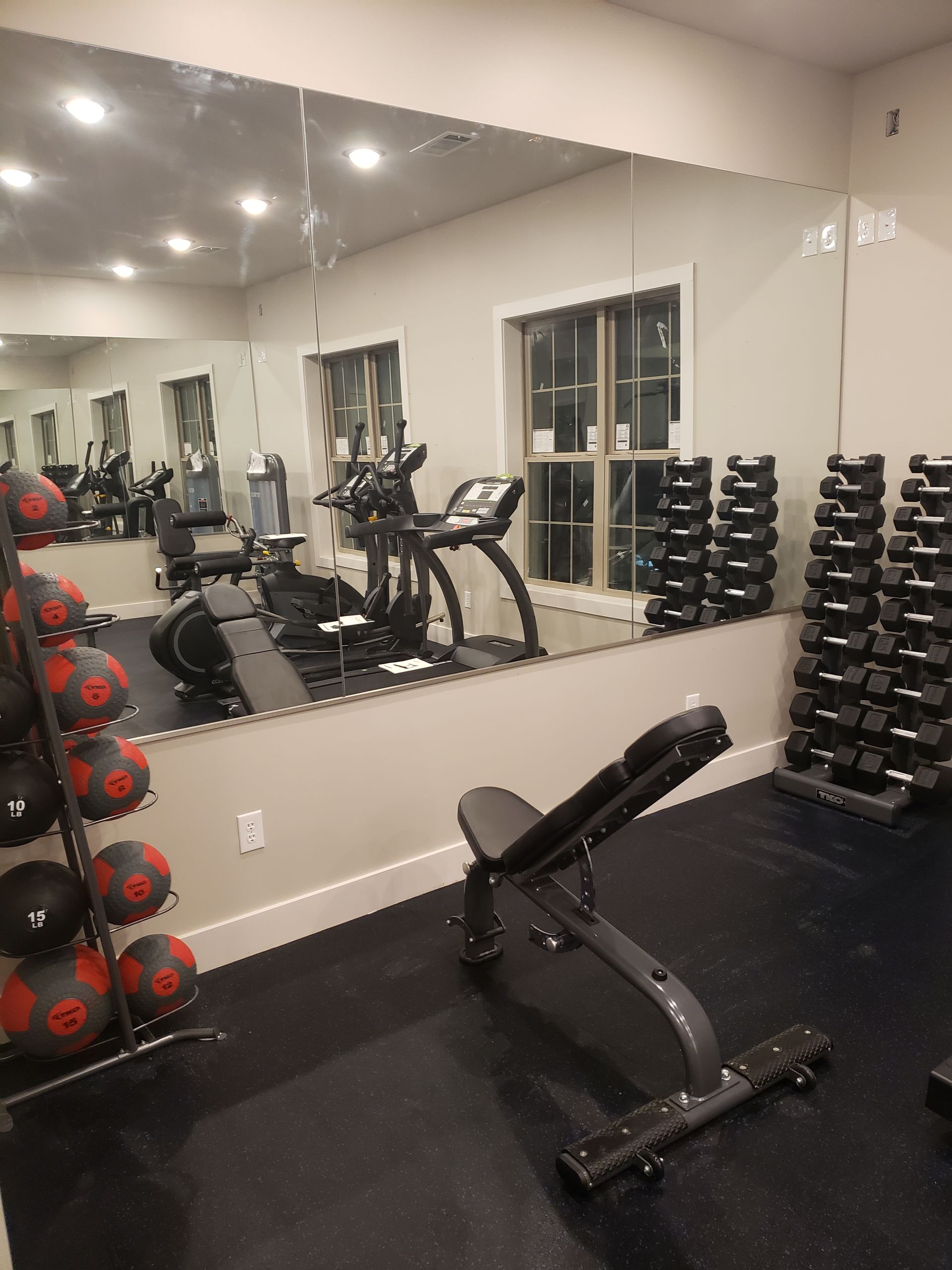 Gym room with mirrored wall, dumbbells, weight bench, and cardio equipment on black flooring