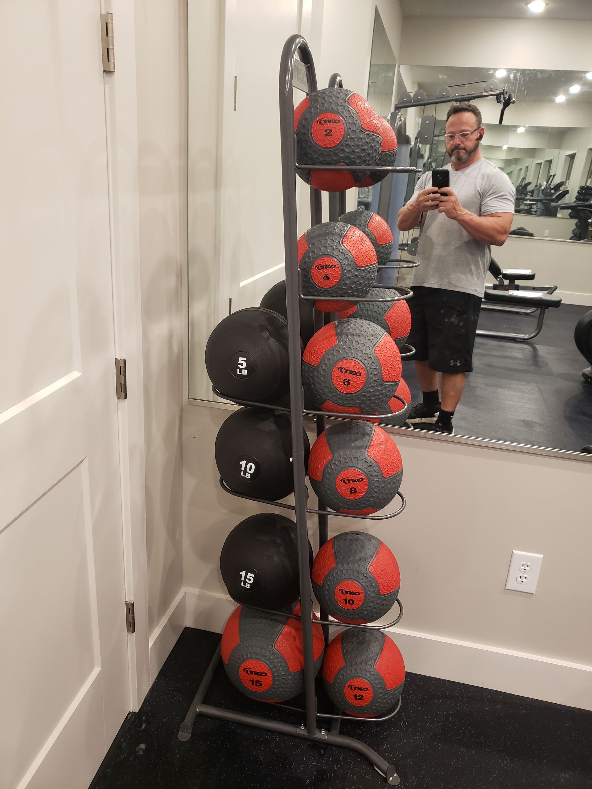 Fitness room with a vertical rack of black-and-red medicine balls beside a mirror and a person taking a selfie.