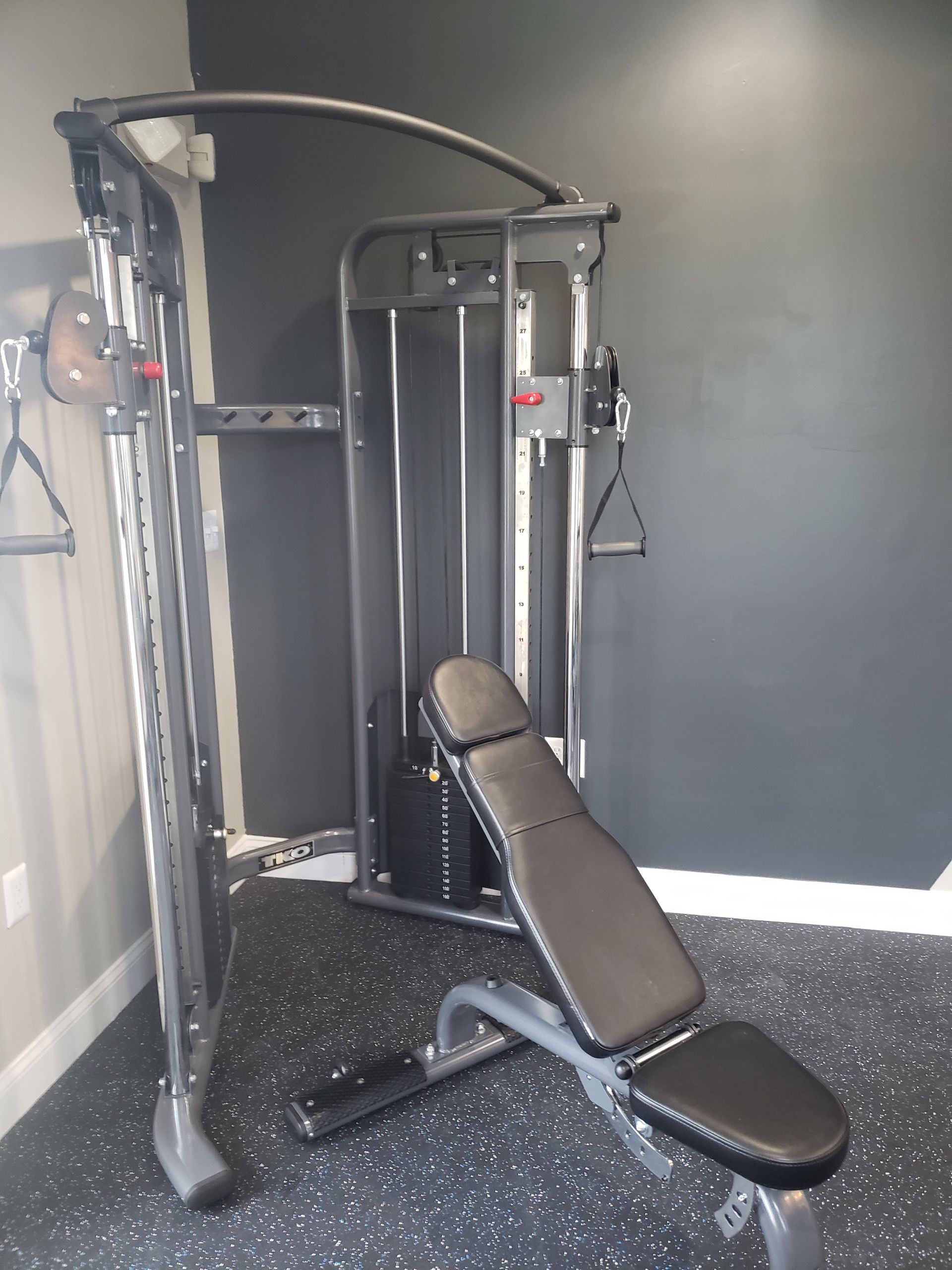 Gym cable machine with adjustable bench in front of a dark gray wall.
