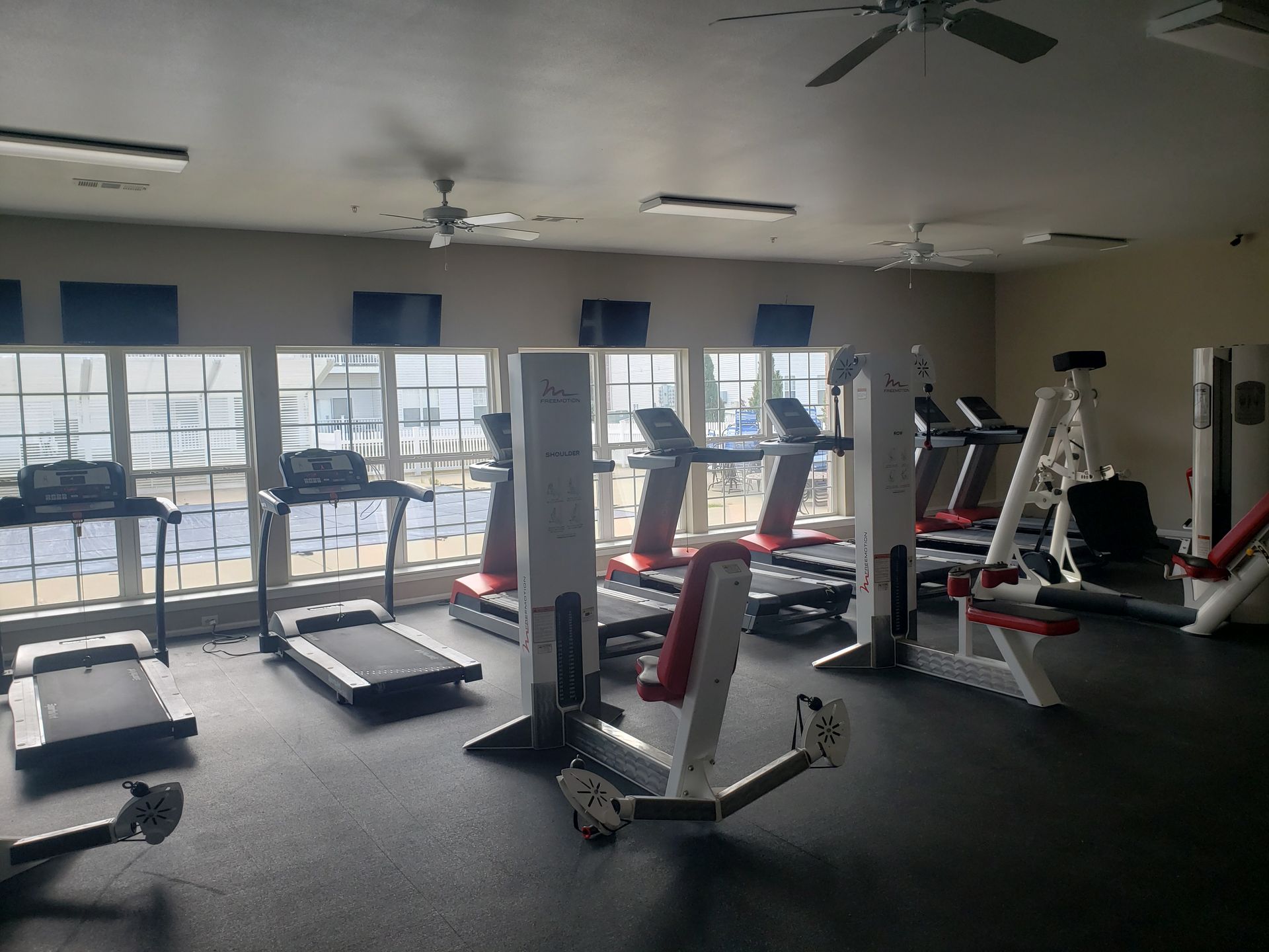 Empty gym with treadmills, exercise bikes, and weight machines near large windows.