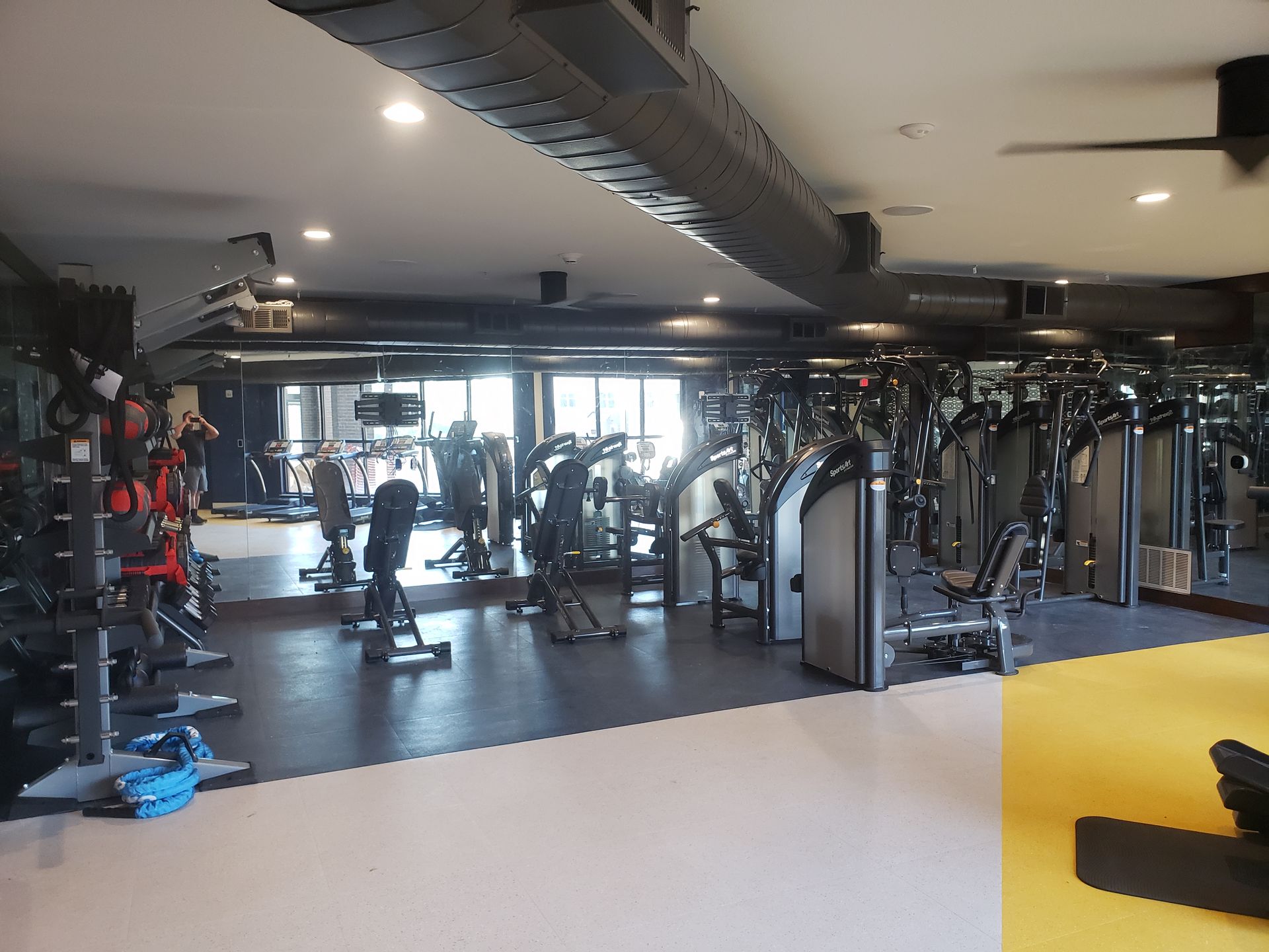Gym interior with cardio machines, weight racks, and a few people exercising near large windows