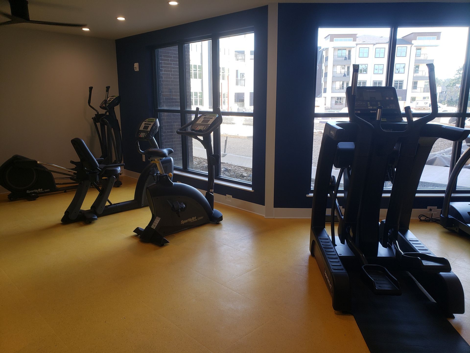Empty gym with exercise machines on a yellow floor beside large windows.