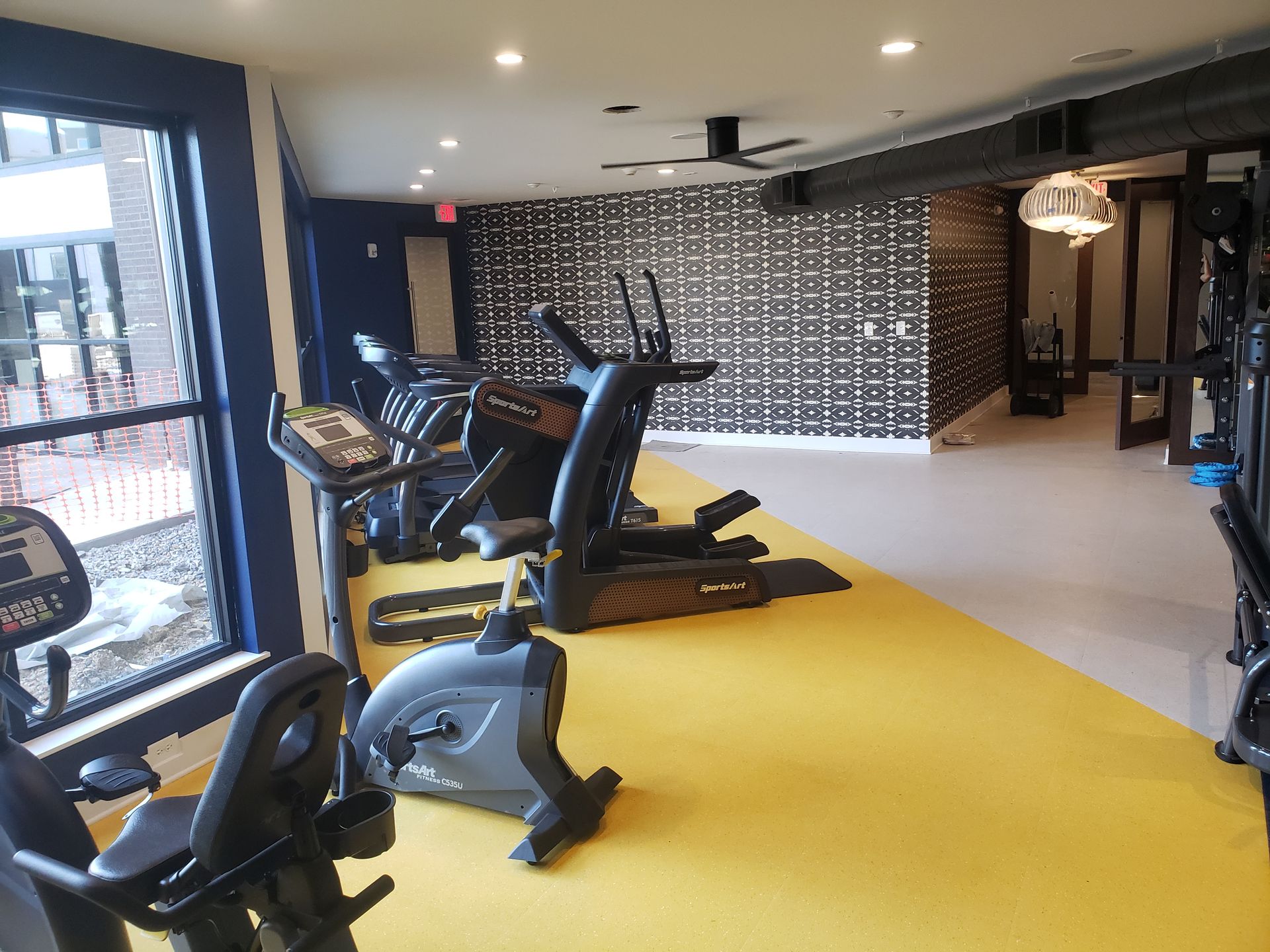 Gym room with treadmills and exercise bikes on a bright yellow floor, beside large windows.