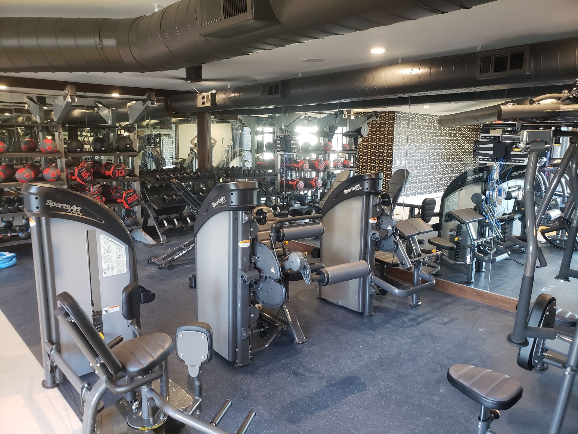 Gym with rows of exercise machines, mirrors, and exposed ductwork in a bright workout room.