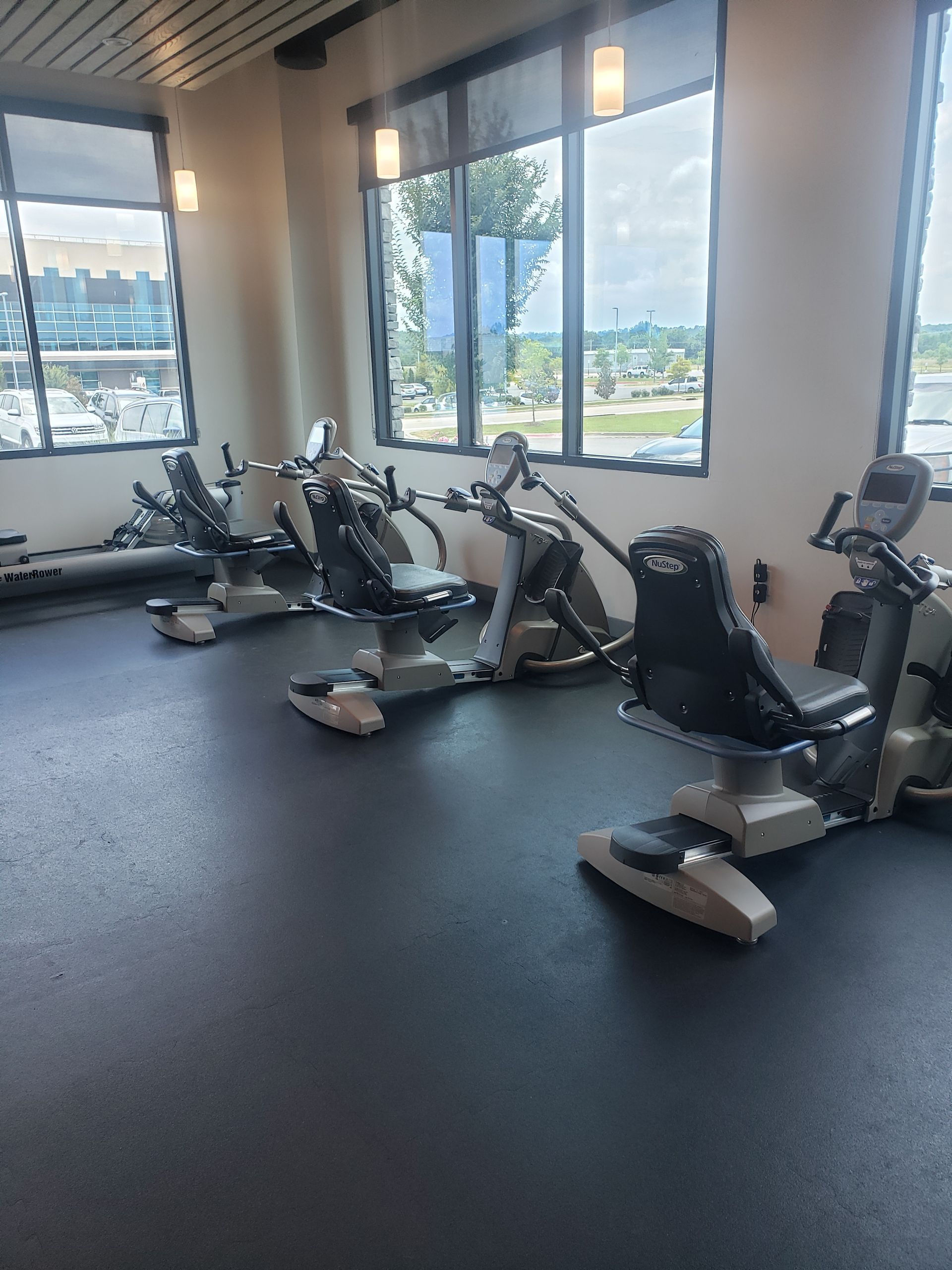Indoor gym with several rowing machines by large windows, overlooking a sunny outdoor view
