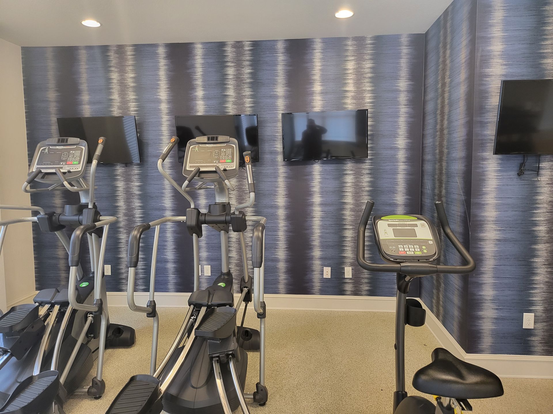 Gym room with treadmills, ellipticals, wall-mounted TVs, and a patterned blue accent wall
