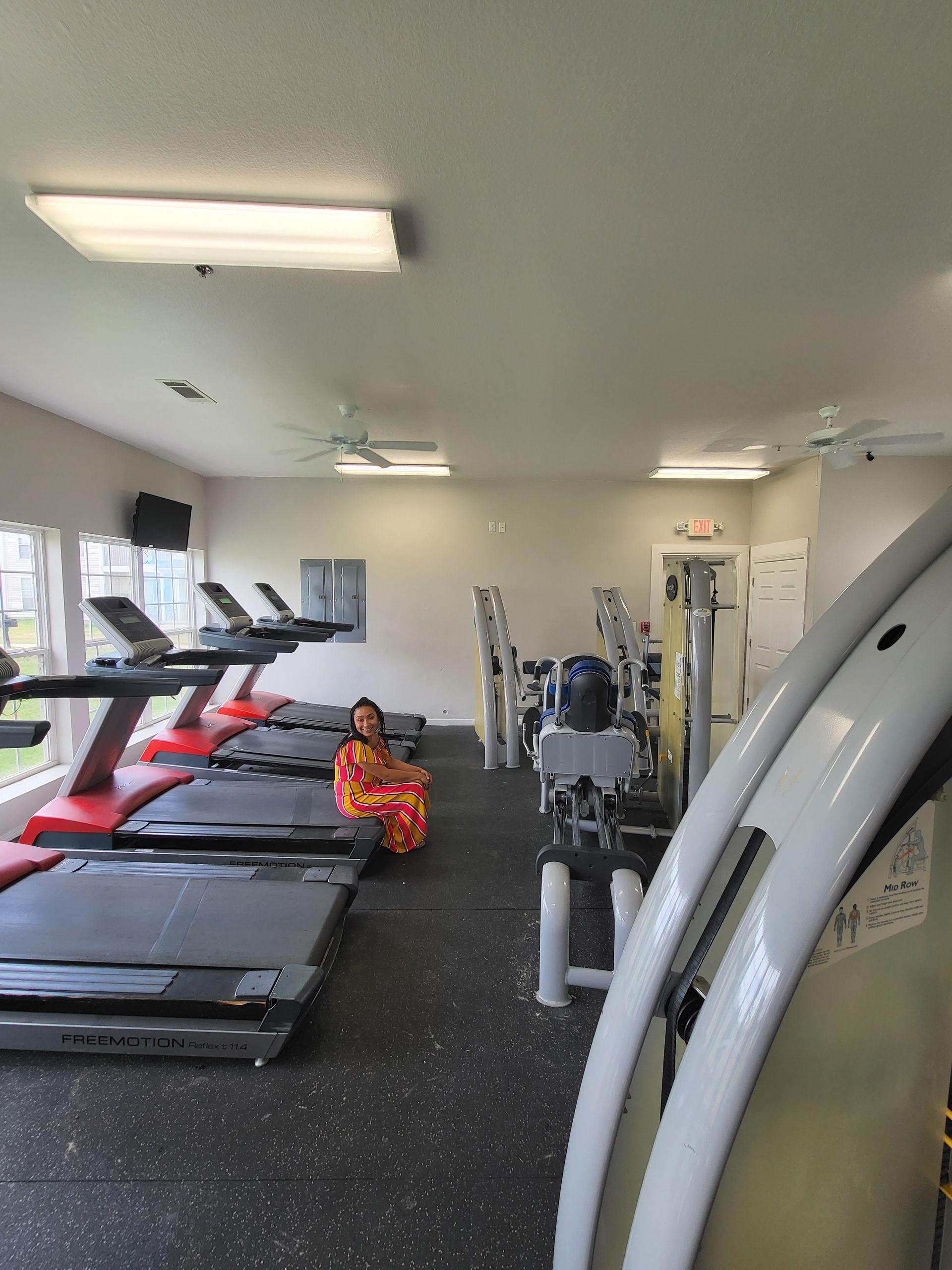 Gym interior with treadmills, weight machines, and a person in a bright orange outfit sitting on the floor