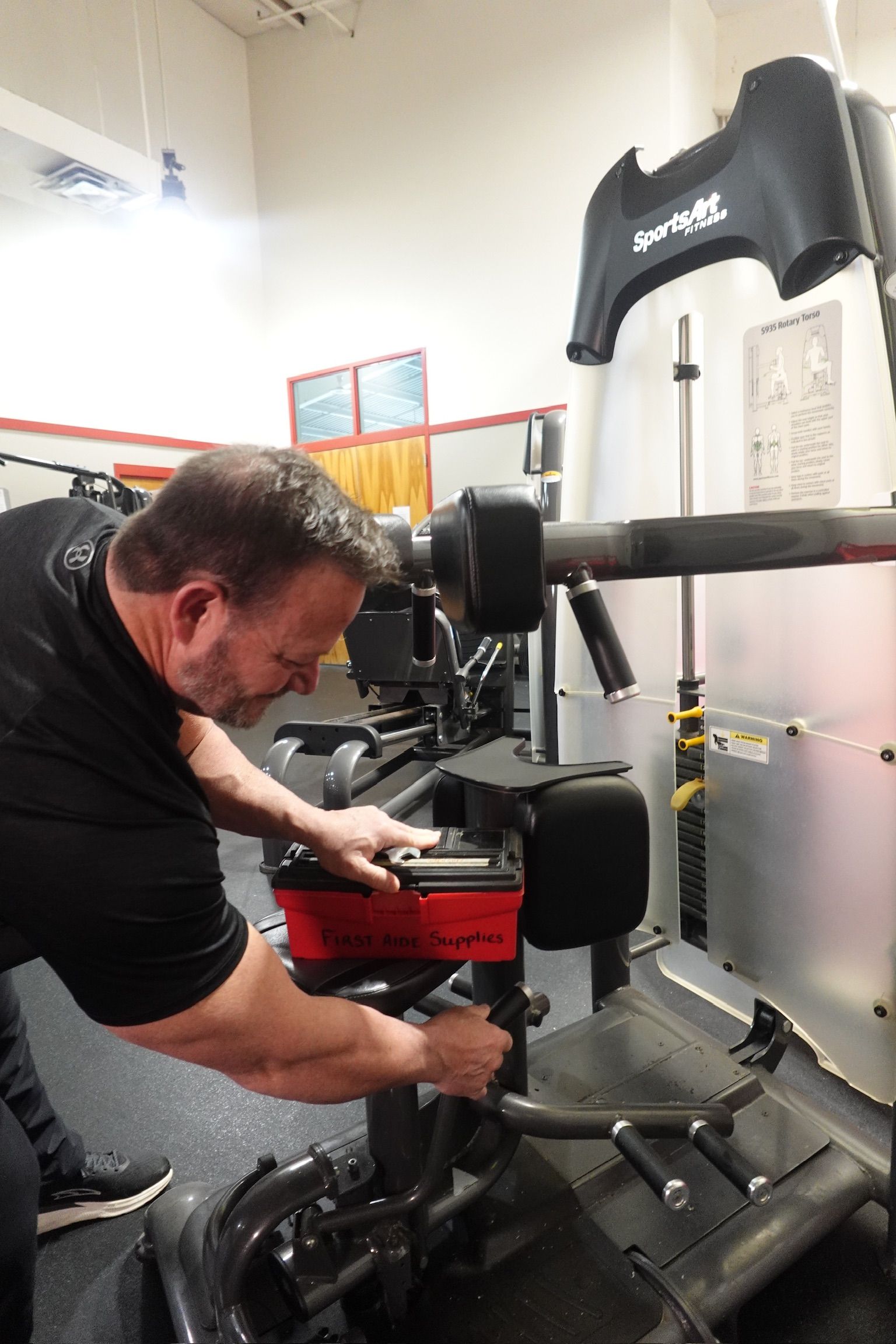 Man adjusting a red and black exercise machine in a gym room with white walls and mirrors