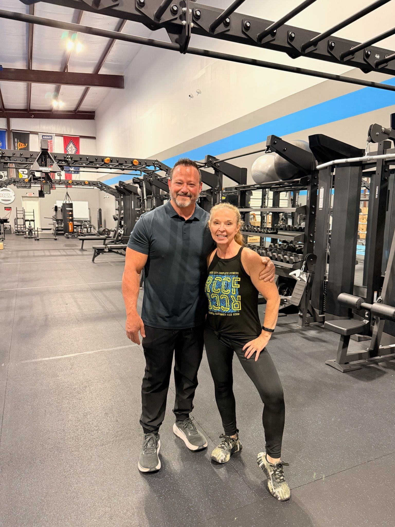Two people posing together in a gym, with exercise machines and equipment in the background.