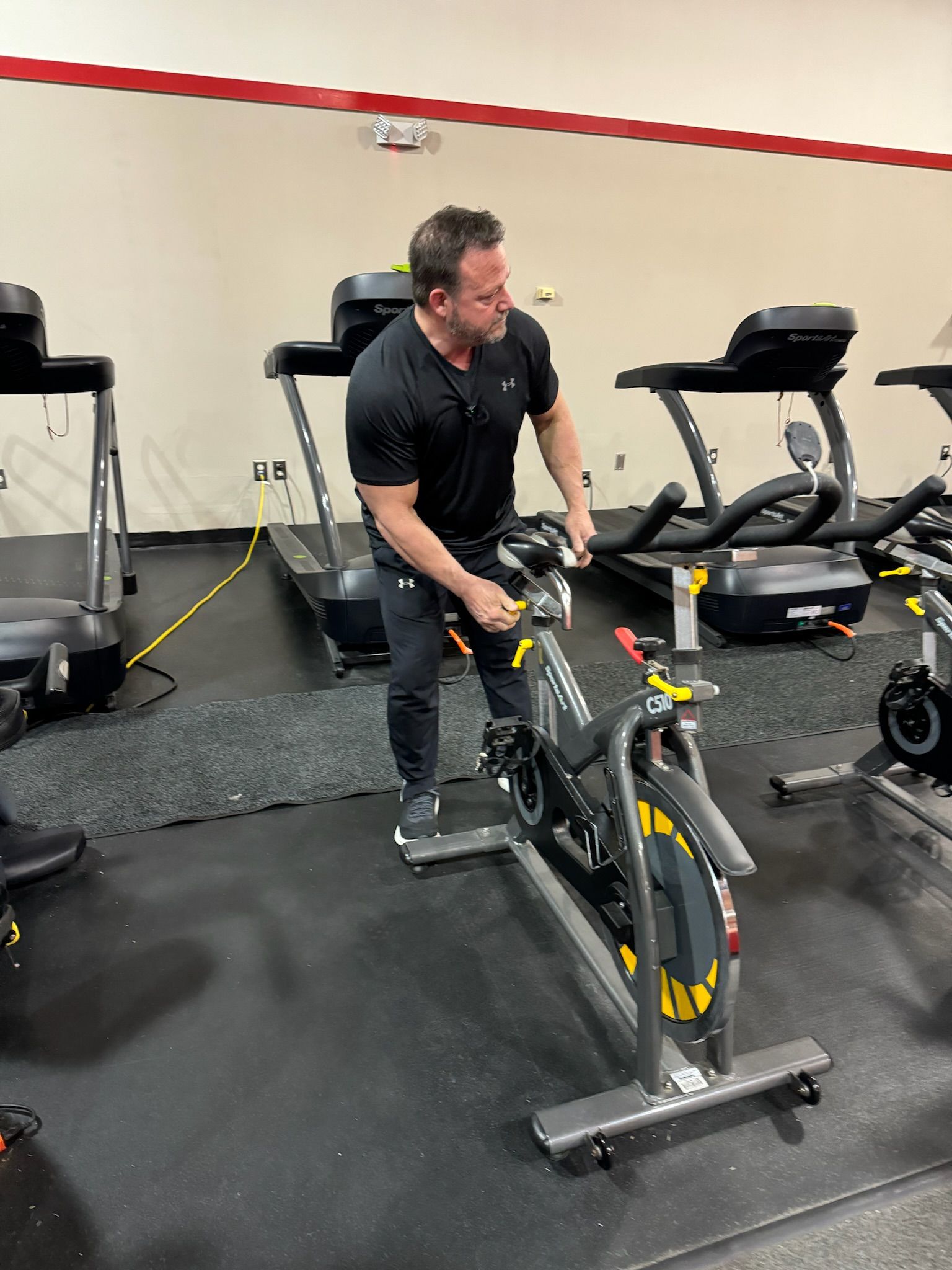 Man adjusting a stationary bike in a gym, with treadmills and exercise equipment nearby.