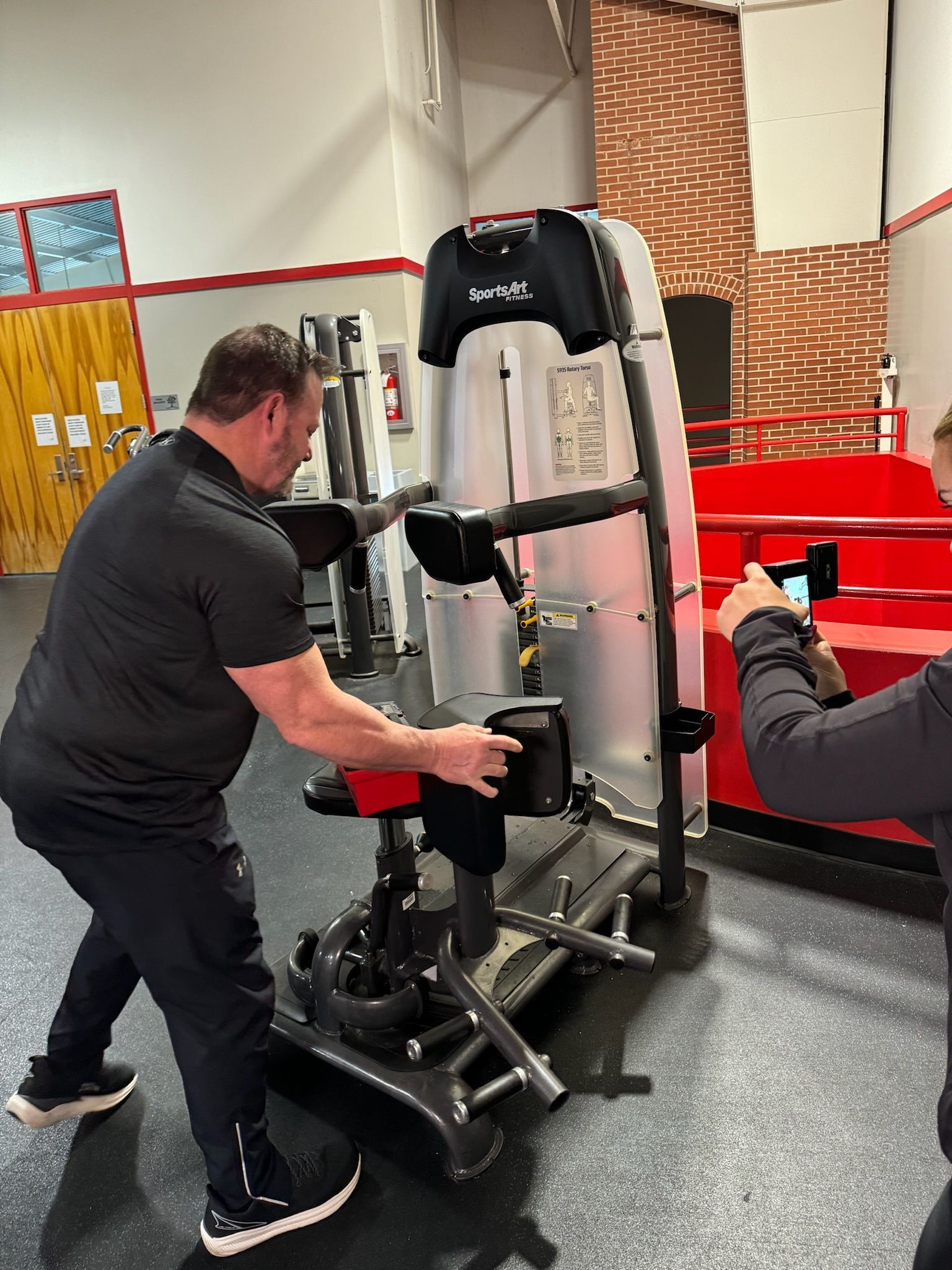Man adjusting a black exercise machine in a gym with red equipment and brick walls.