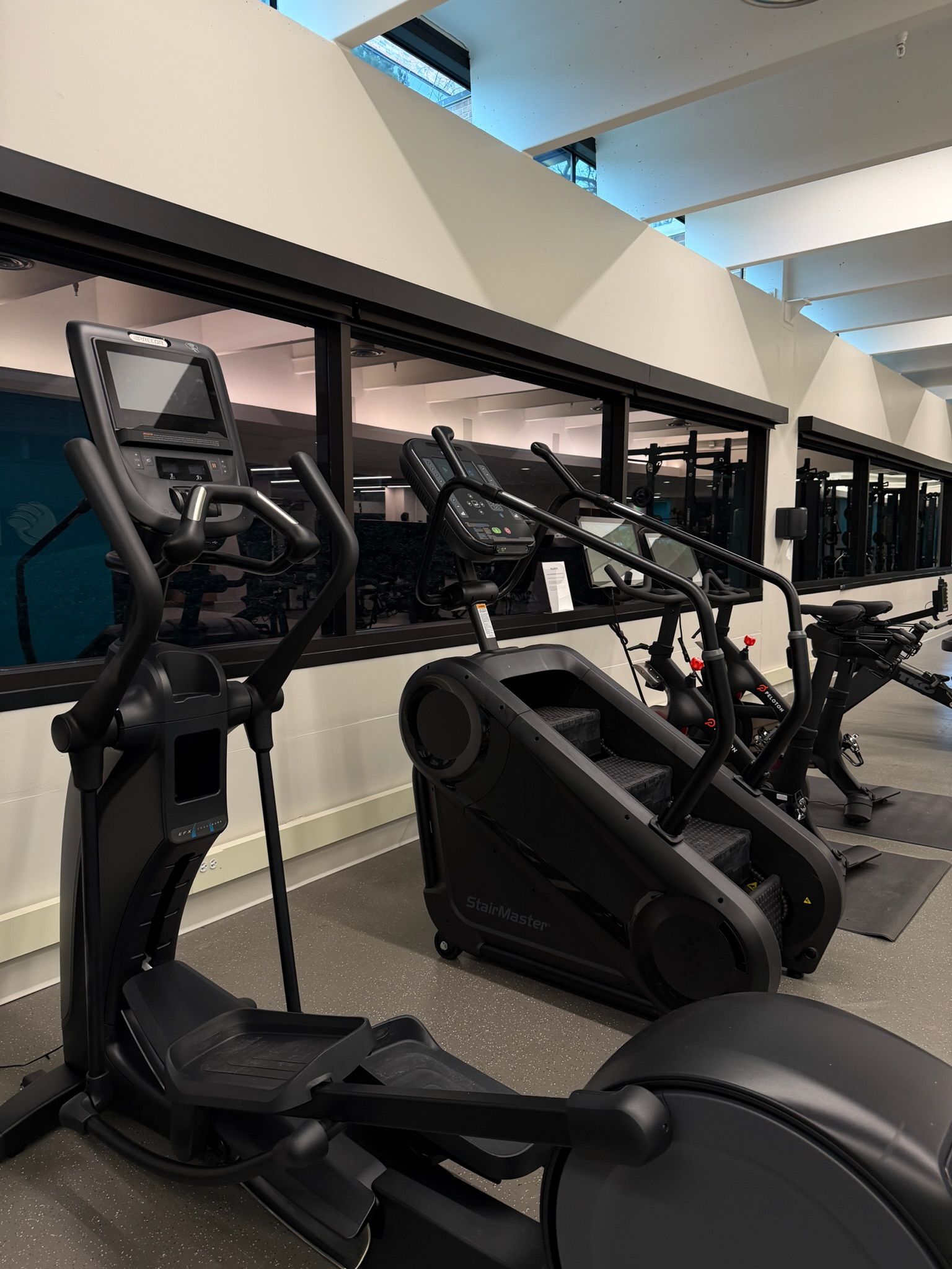 Row of ellipticals in a gym beside a mirrored wall and windowed ceiling