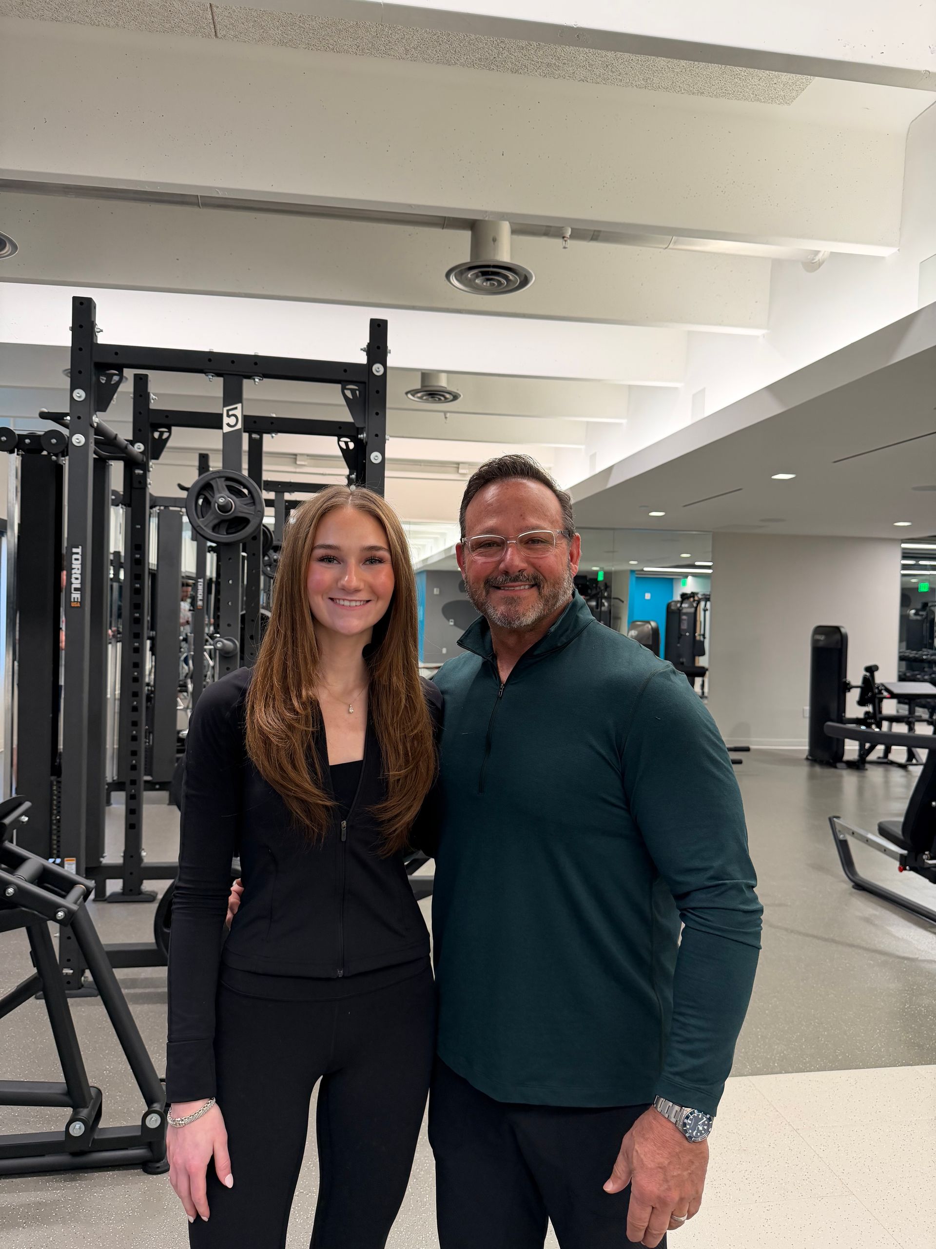 Two people posing in a gym beside exercise equipment, smiling at the camera.