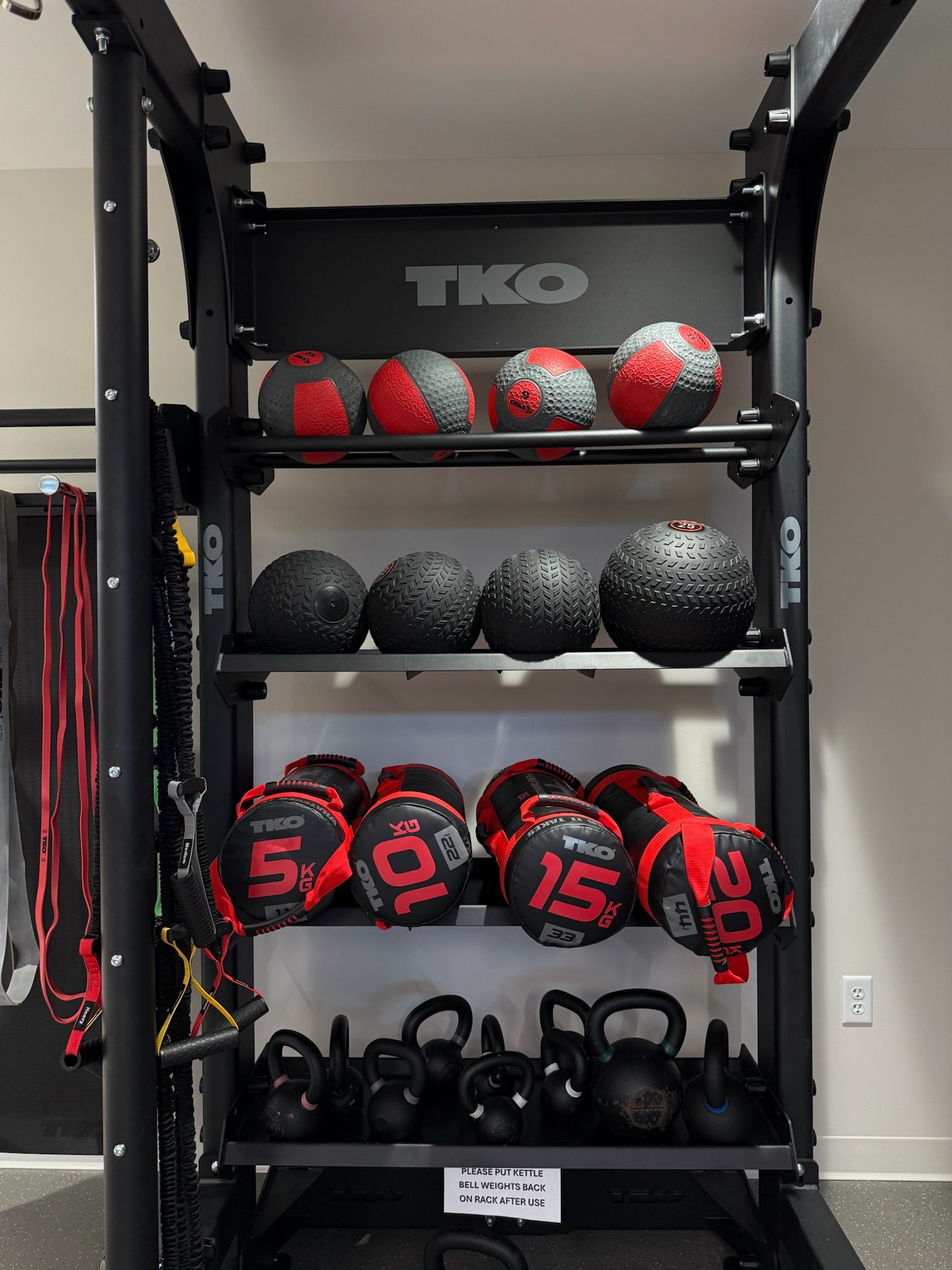 TKO-branded rack with medicine balls, weighted balls, and kettlebells in a gym corner
