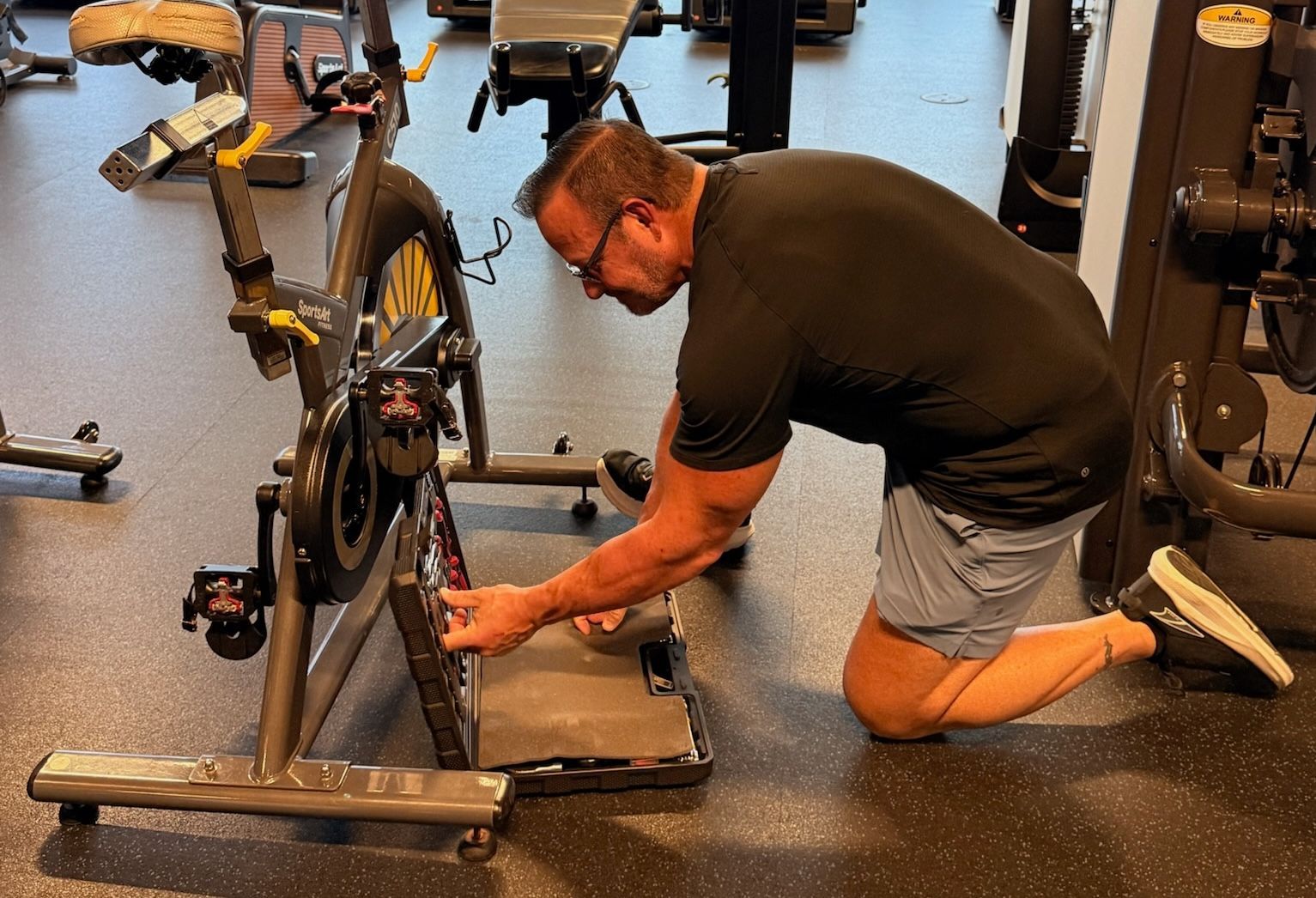 Man kneeling beside a bicycle, adjusting the rear wheel in a gym-like room.
