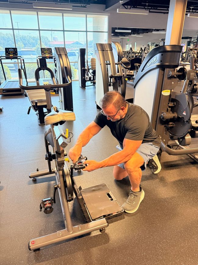 Man kneeling beside a bicycle, adjusting the rear wheel in a gym-like room.