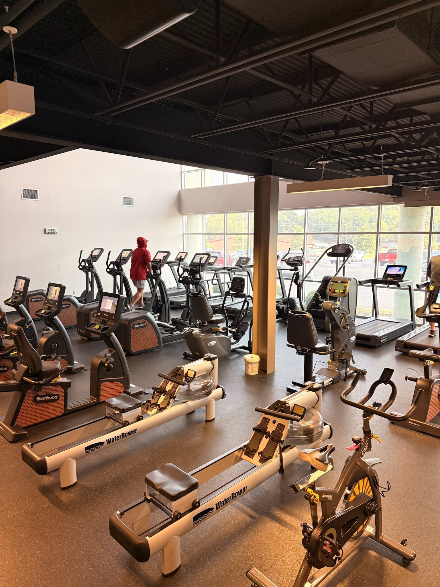 Indoor gym with rows of stationary bikes and treadmills under a dark ceiling, large windows on the right.