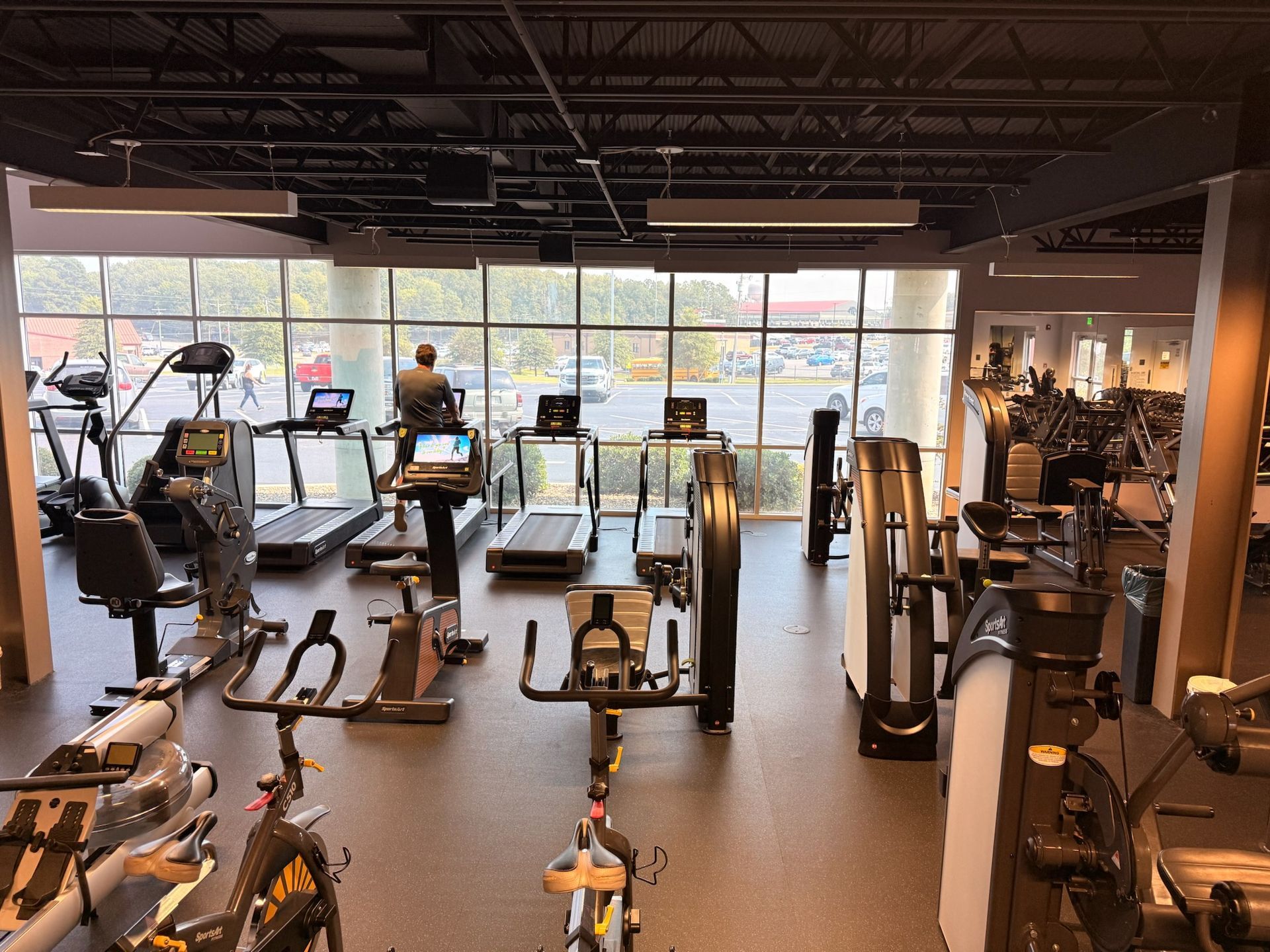 Modern gym interior with treadmills, exercise bikes, weight machines, and large windows showing daylight