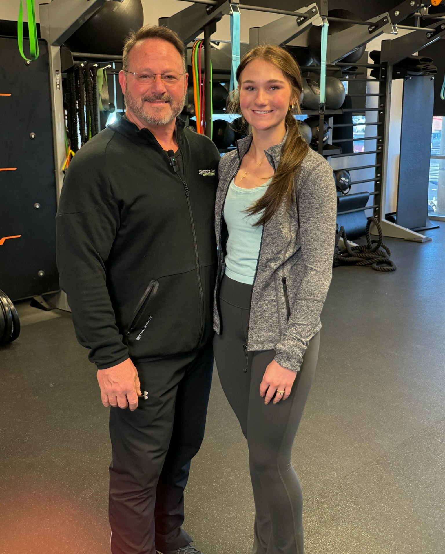 Two people posing together in a gym, wearing black workout clothes.