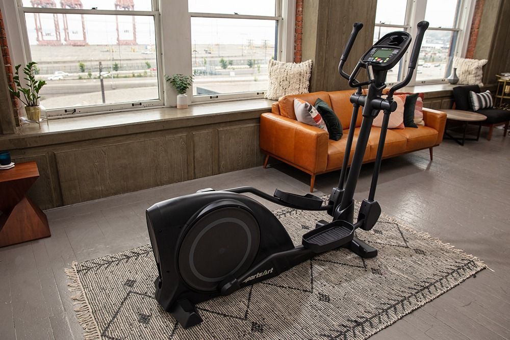 Elliptical machine on a rug in a bright living room with a sofa and large windows