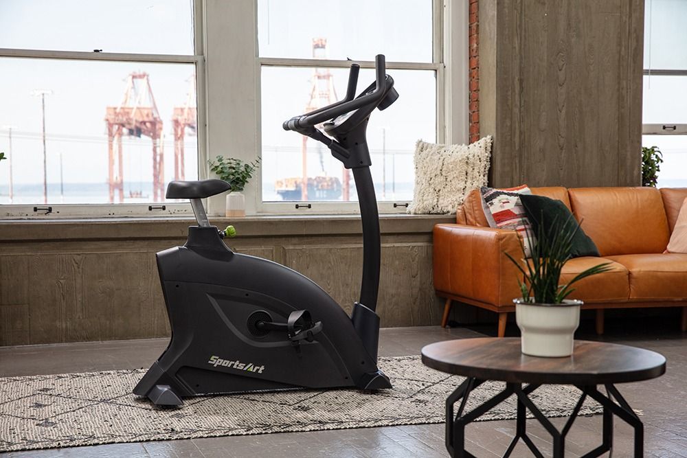 Black stationary exercise bike in a bright living room beside an orange sofa and coffee table.