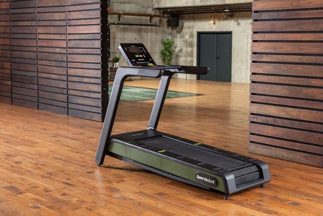 Treadmill in a wooden-floored gym with wall-mounted slats and a dark doorway in the background
