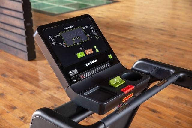 Treadmill in a wooden-floored gym with wall-mounted slats and a dark doorway in the background