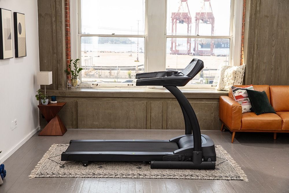 Treadmill in a bright living room beside a couch and large window.