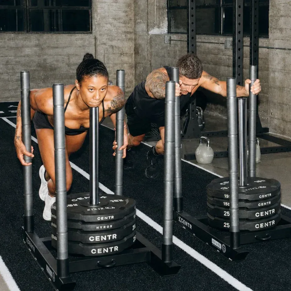 Two athletes pushing weighted sleds in a gym, leaning forward on a black rubber floor.