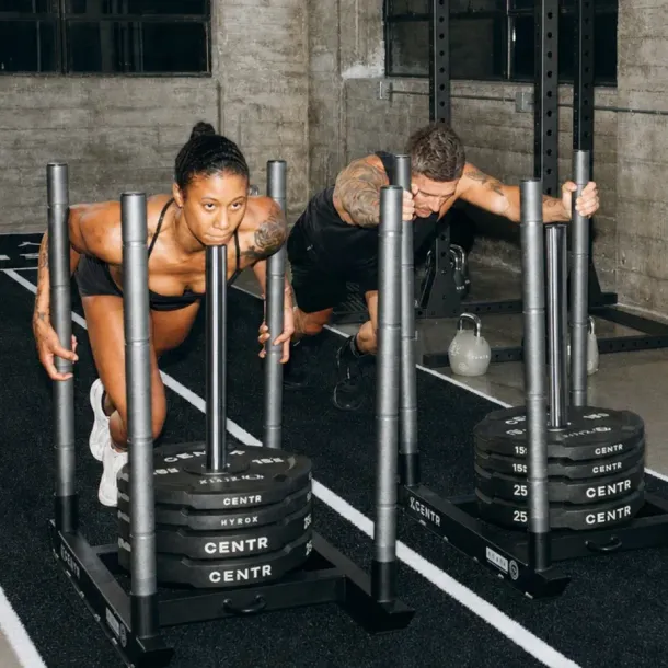 Two athletes pushing weighted sleds in a gym, leaning forward on a black rubber floor.