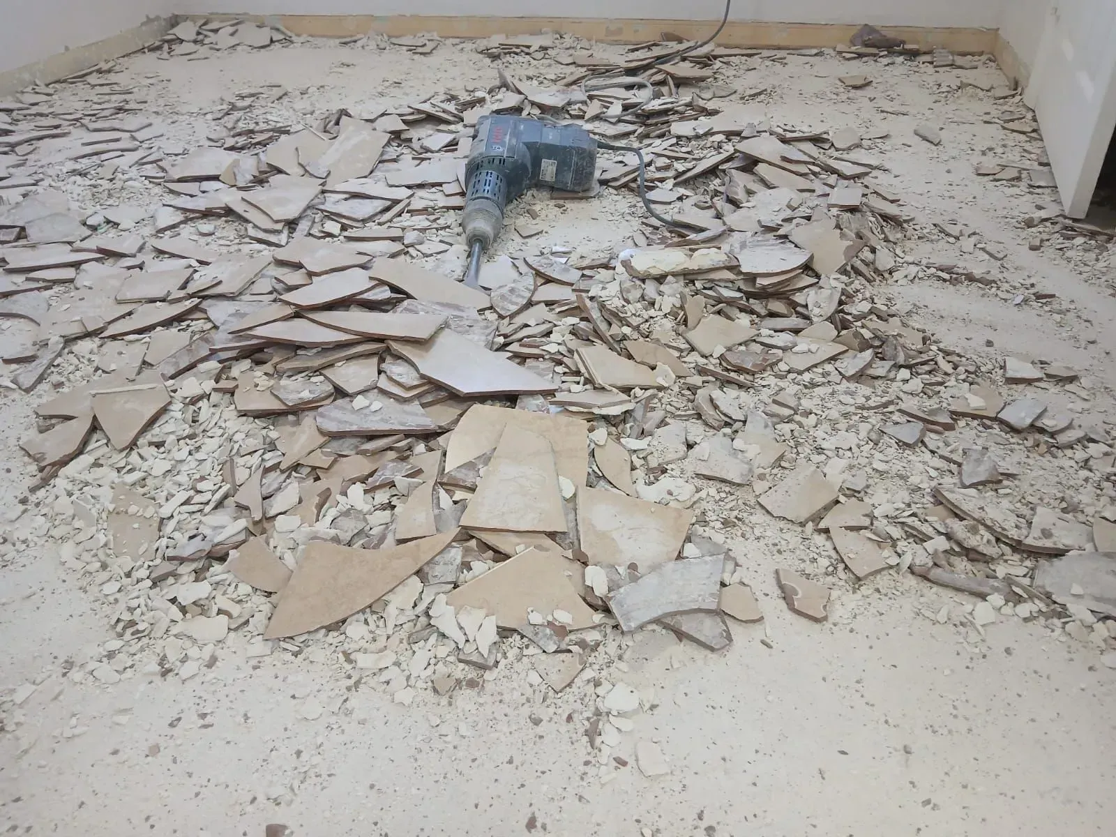 Crumbling beige floor tiles and debris scattered across a room, with a blue power tool near the back wall