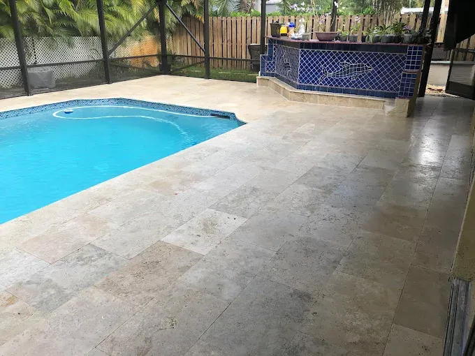 Poolside patio with light stone tiles beside a blue swimming pool and a tiled outdoor bar area