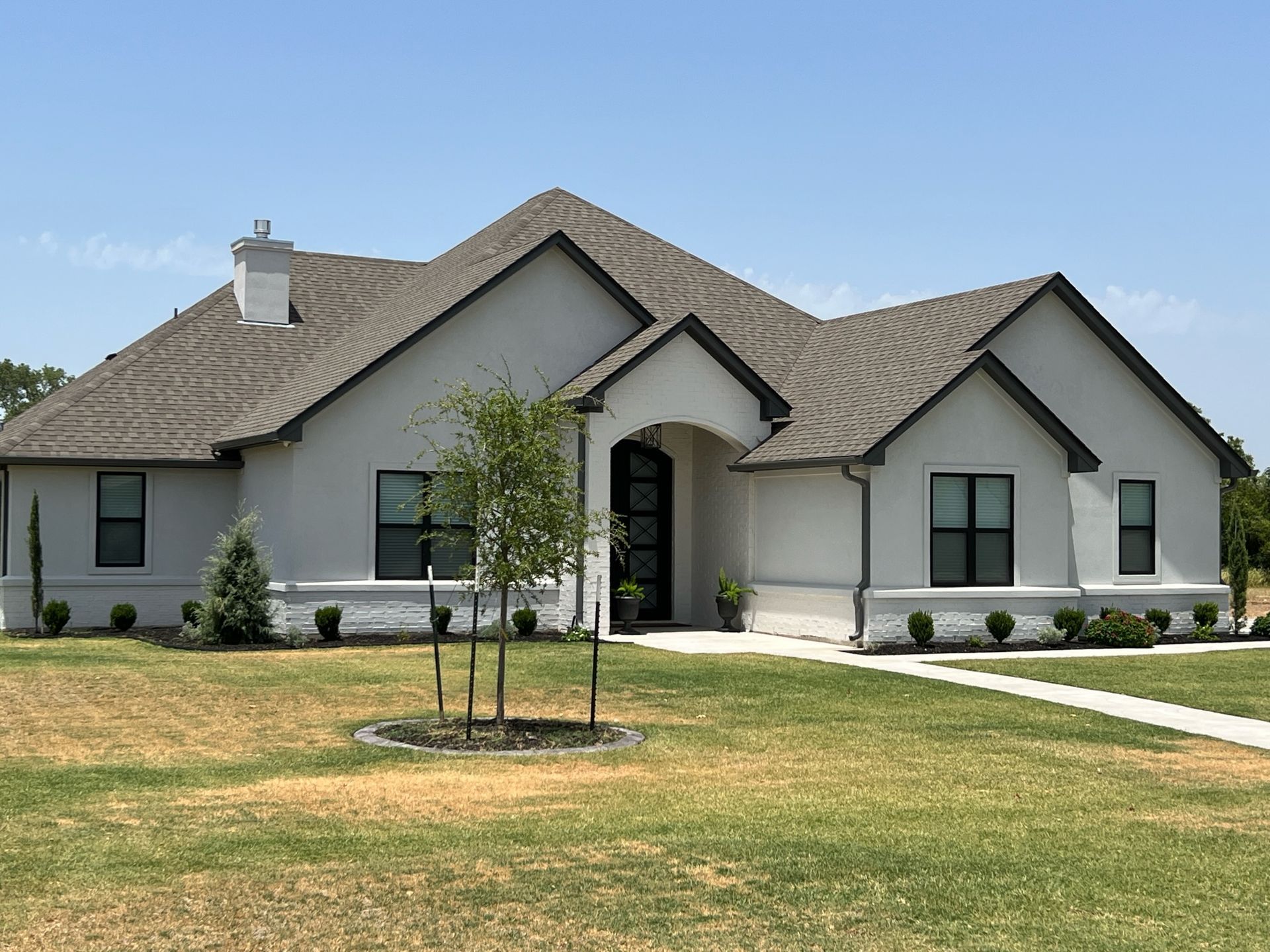 Single-story white house with dark roof, front porch, and young trees in a sunny yard.